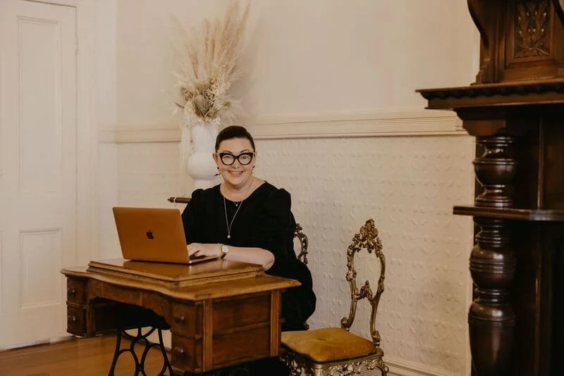 A woman with glasses and dark hair sitting at a wooden desk with an open MacBook, smiling in a warmly decorated room with a white wall, a large vase with dried plants, and vintage furniture.