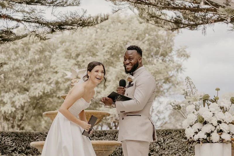 A bride and groom are smiling during their wedding ceremony outdoors, with the officiant holding a microphone and a script. The backdrop includes trees and floral arrangements.