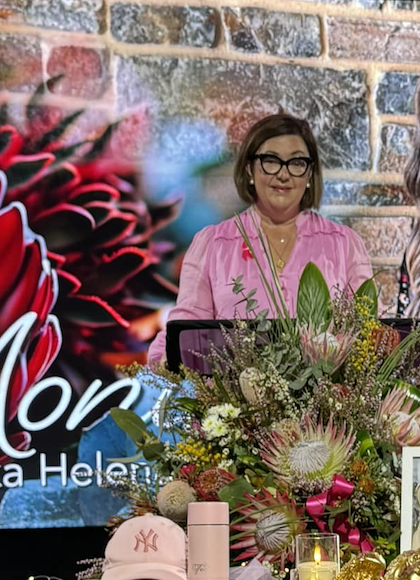 A woman with short brown hair, wearing glasses and a pink shirt, standing behind a table with a large bouquet of flowers at an indoor event or presentation.