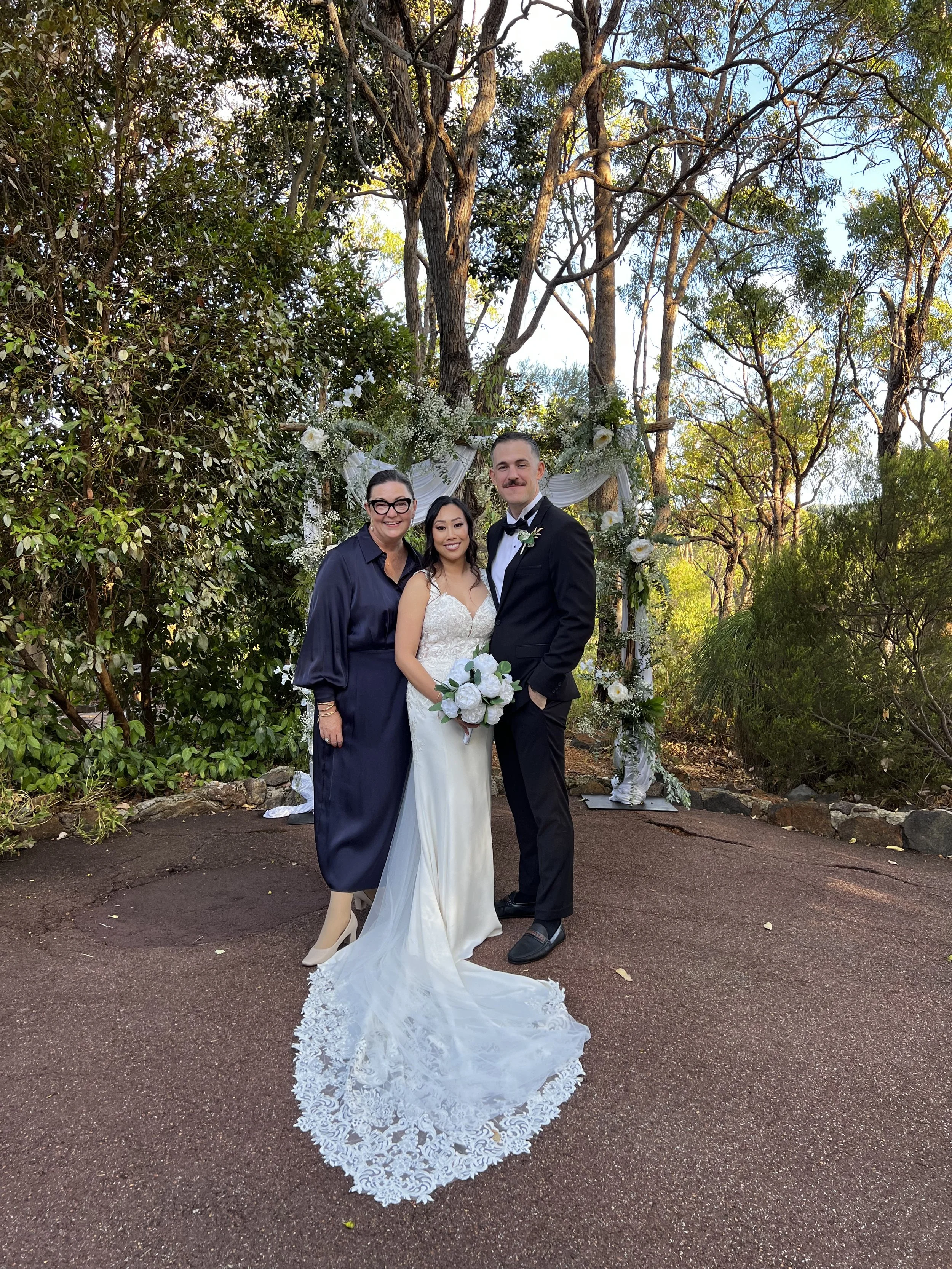 A bride and groom standing outdoors with two women, surrounded by trees and greenery, at a wedding ceremony setting with floral decorations in the background.