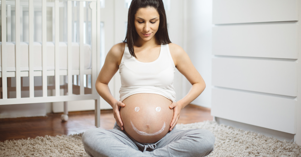Pregnant woman sitting cross-legged on a rug, with white markings on her belly, in a bright bedroom.