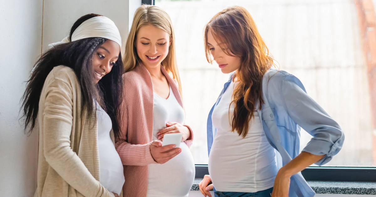 Three pregnant women standing together in front of a window, smiling and looking at a phone.