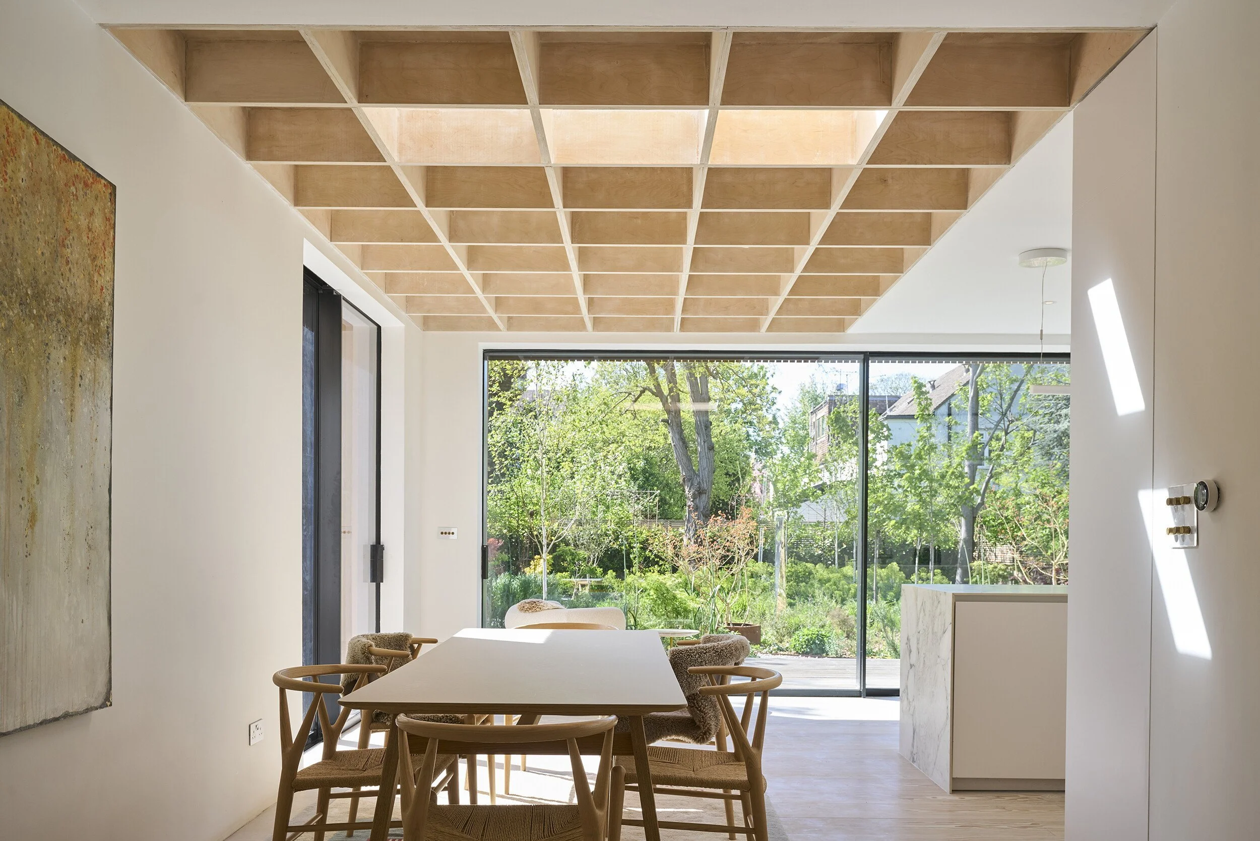 Modern dining area with a wooden table and chairs, large windows revealing a green outdoor space, and a ceiling with a grid pattern of wooden squares.