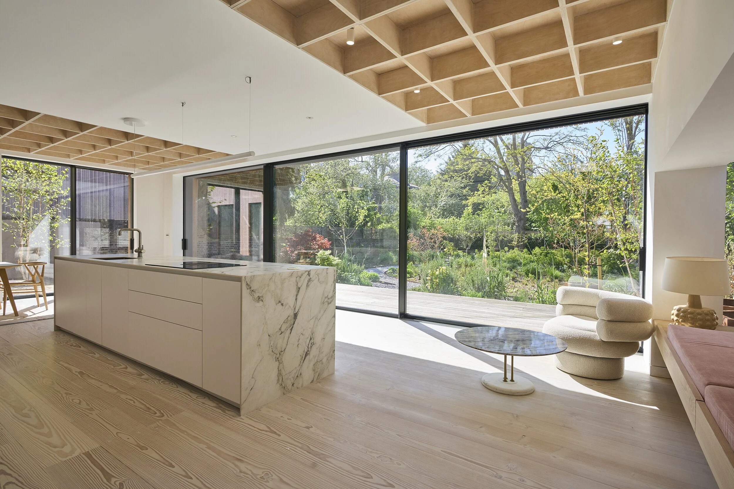 Modern kitchen and living room with large glass doors overlooking a garden, featuring light wood flooring, a marble kitchen island, and built in banquette seating.