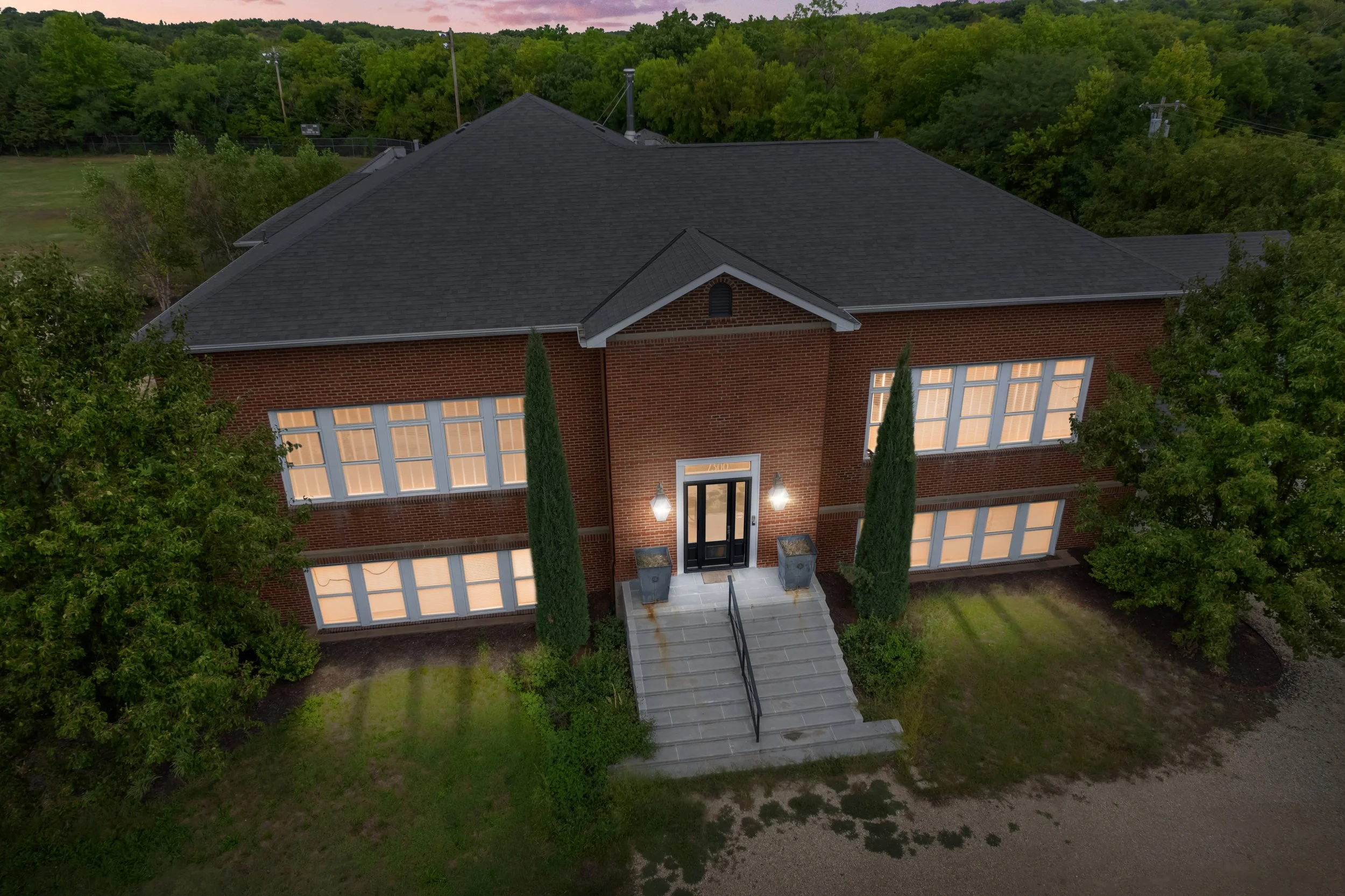 Aerial view of a two-story brick house with a dark roof, illuminated exterior lights, two tall cypress trees flanking the front door, and large windows with warm interior lighting. The house is surrounded by trees and a grassy yard with a gravel driv