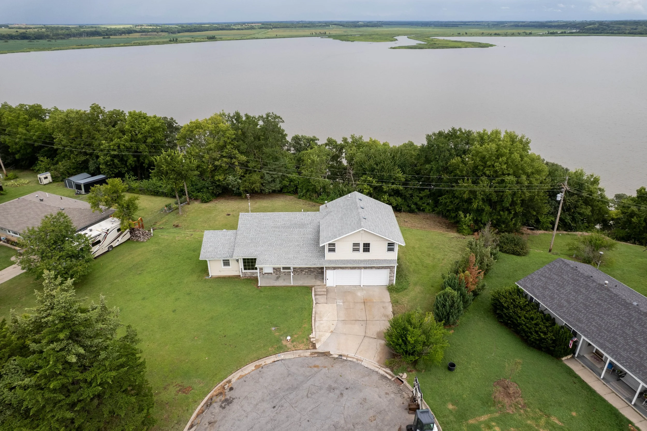 A two-story house with an attached garage, surrounded by a lawn and trees, located near a large body of water. Other houses are nearby.