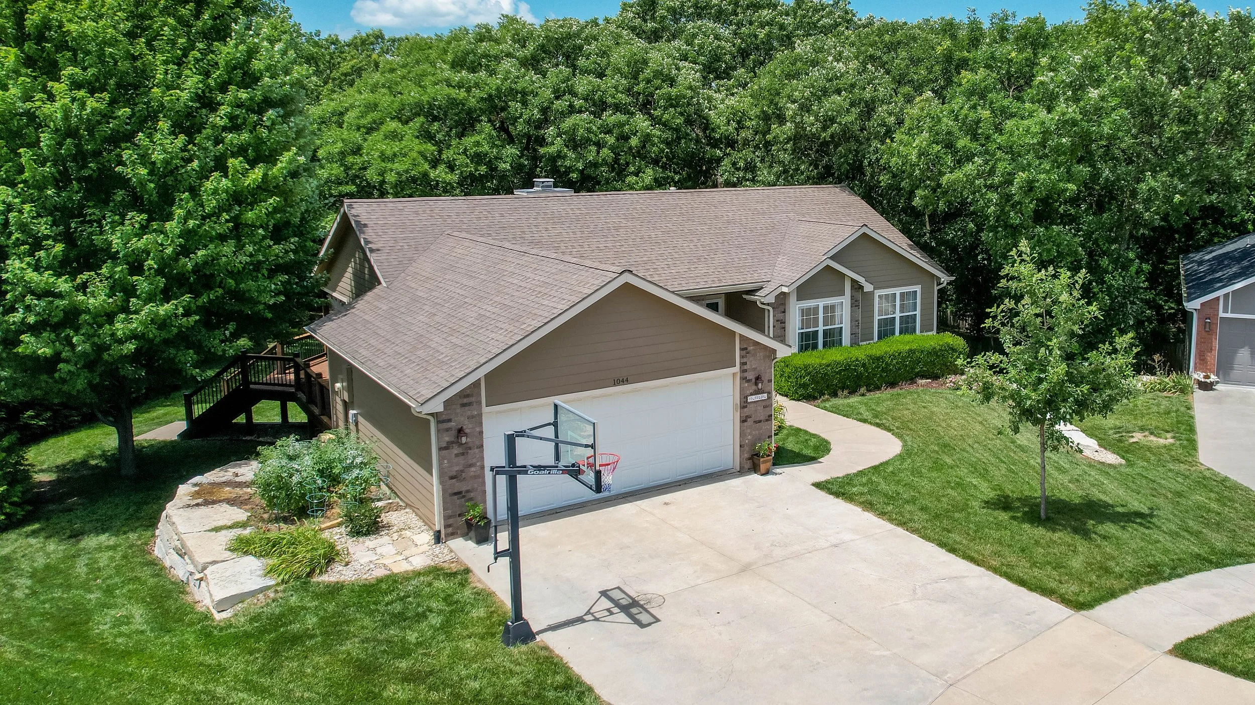 Aerial view of a suburban house with a basketball hoop in the driveway, surrounded by green trees and landscaped yard.