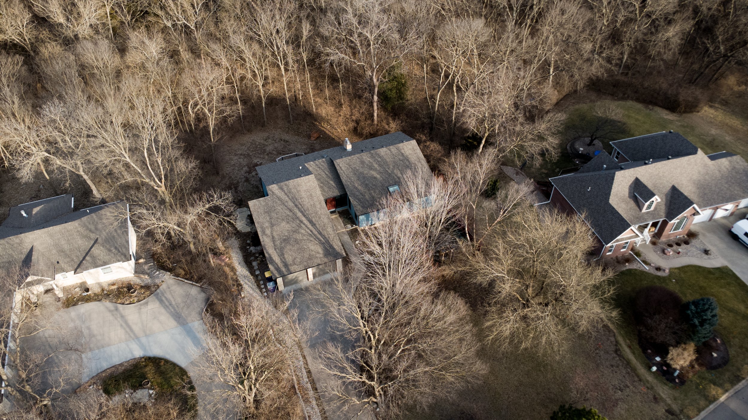 An aerial view of residential houses with trees around, some with bare branches, and a yard with a driveway.