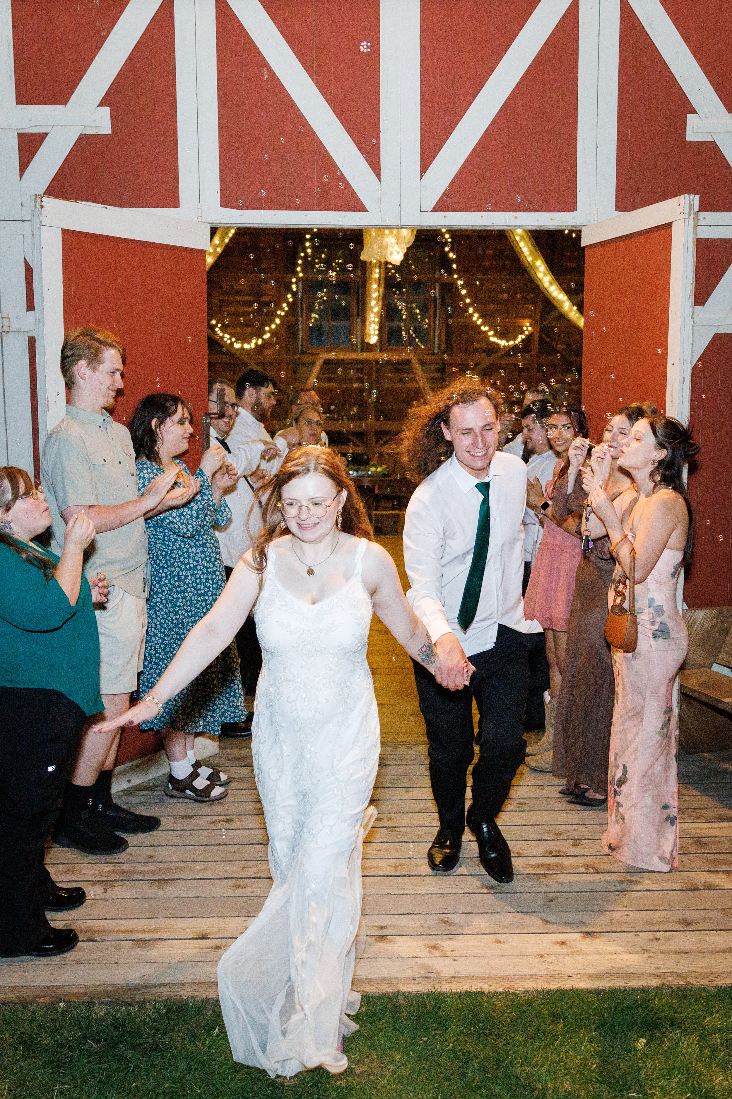 bride and groom running out of red barn door during a bubble exit at their wedding with wedding guest all around