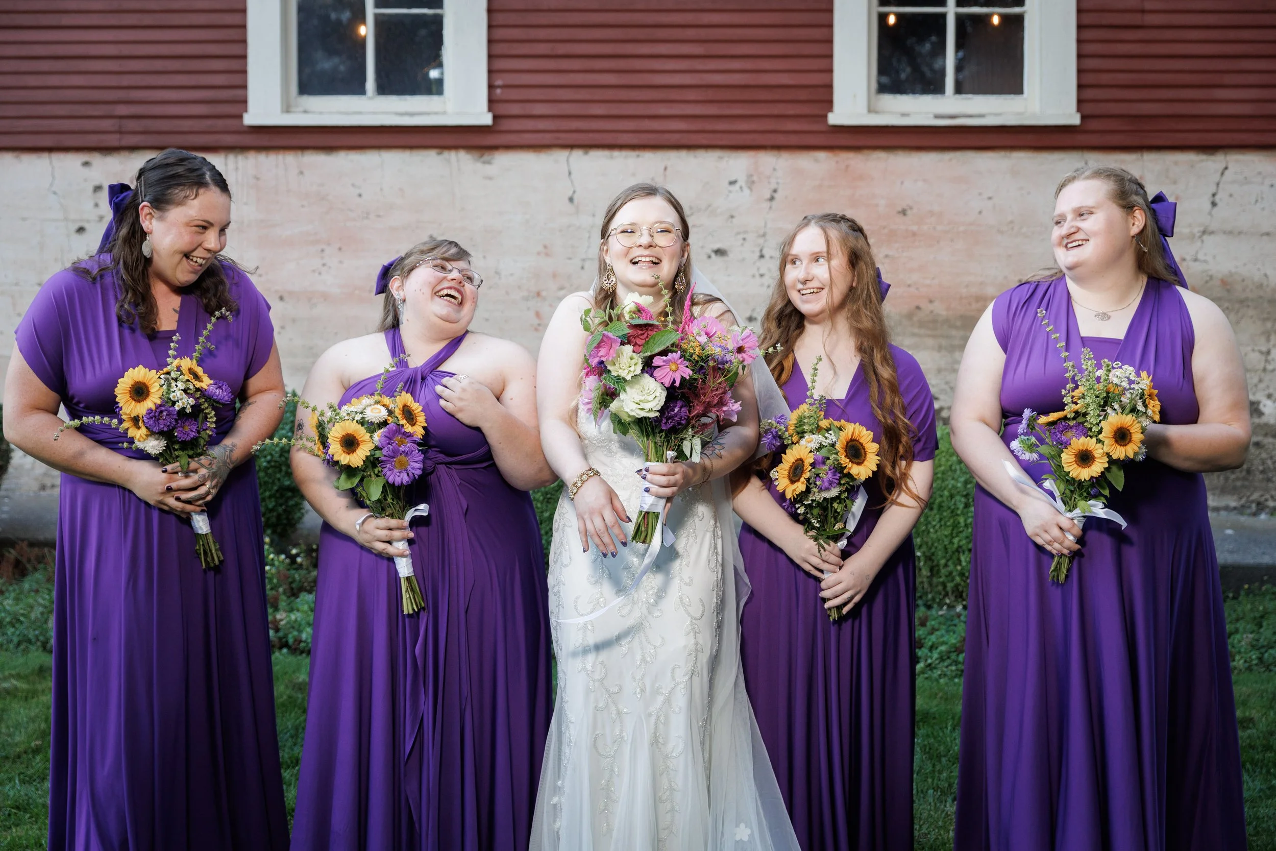 bridesmaids laughing around the bride in front of a red barn during group photos