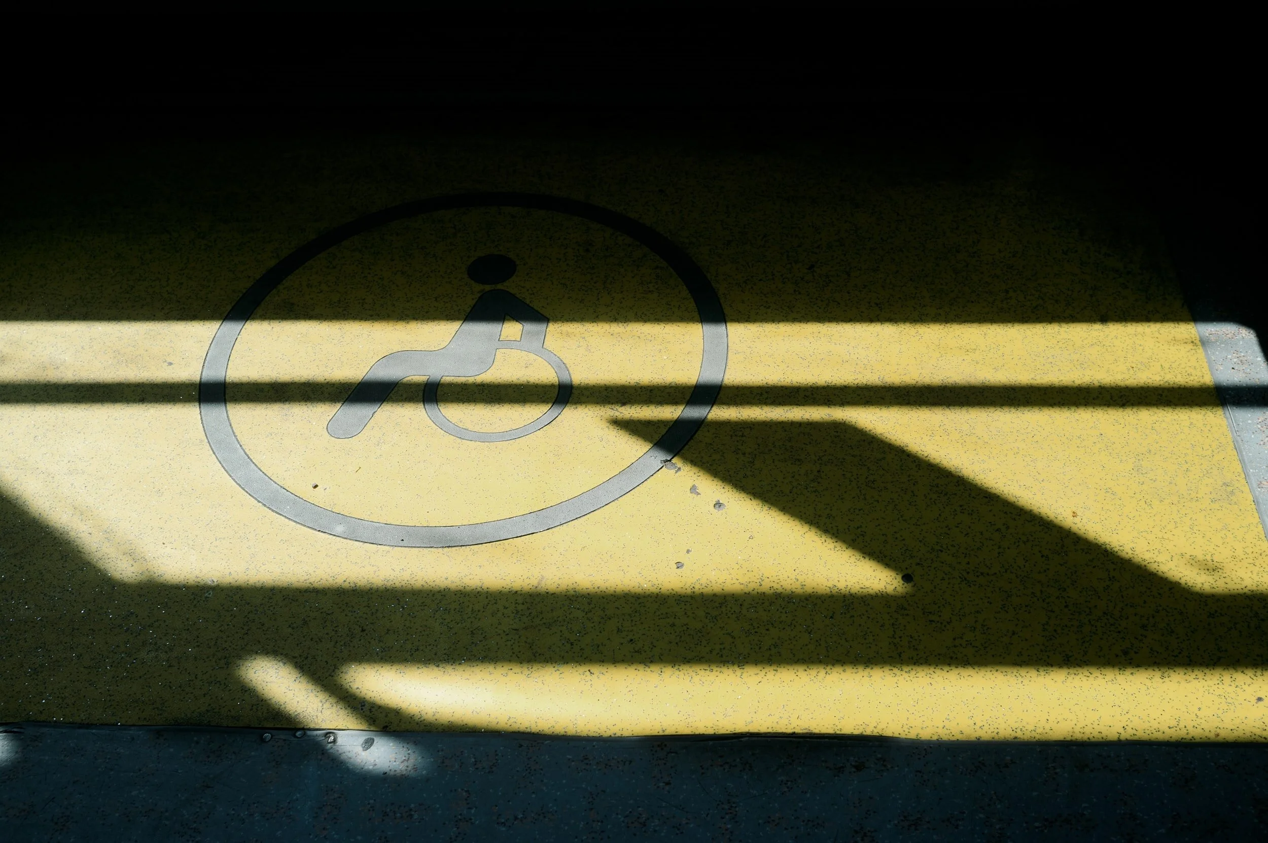 a stock photo of a disabled parking spot with the disabled sign visible in yellow. Shadows are cast over the sign which slightly obscures it.