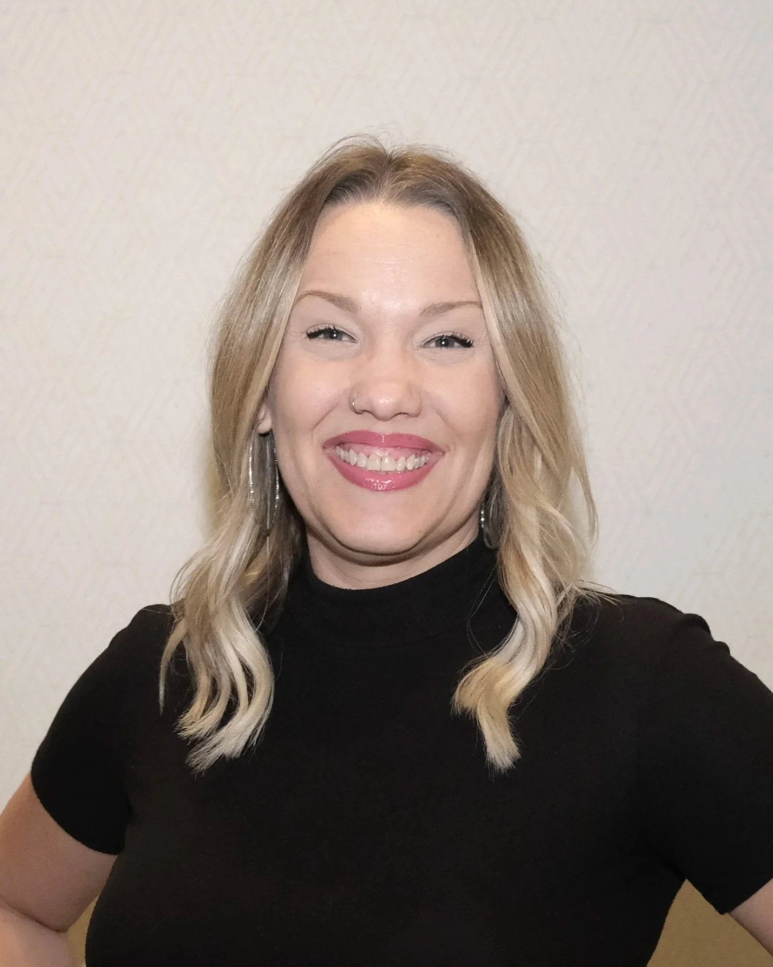 A woman with blonde, shoulder-length hair, smiling outdoors with blurred trees in the background.
