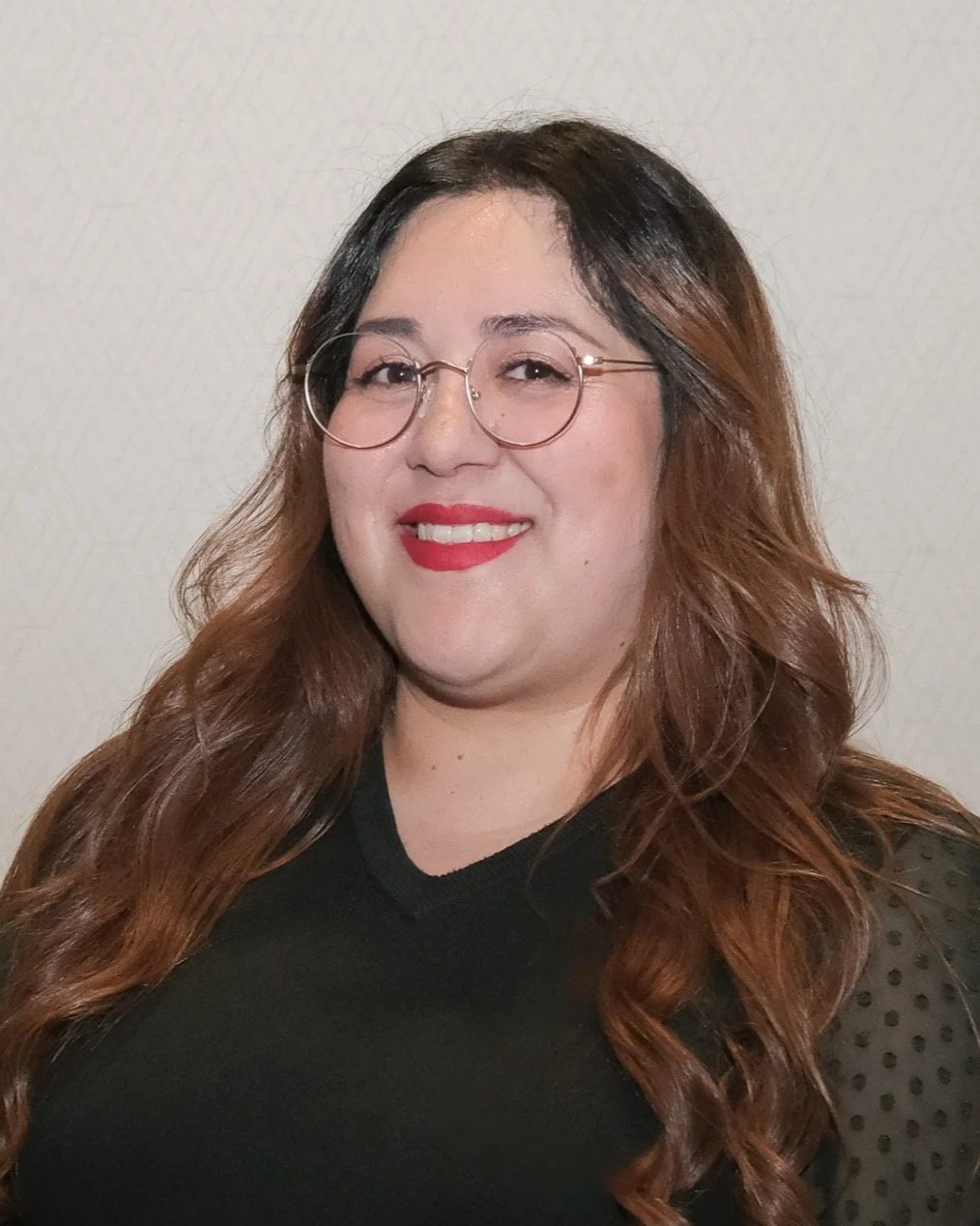 A woman with long wavy hair and glasses smiling outdoors against a wooden fence with trees in the background.