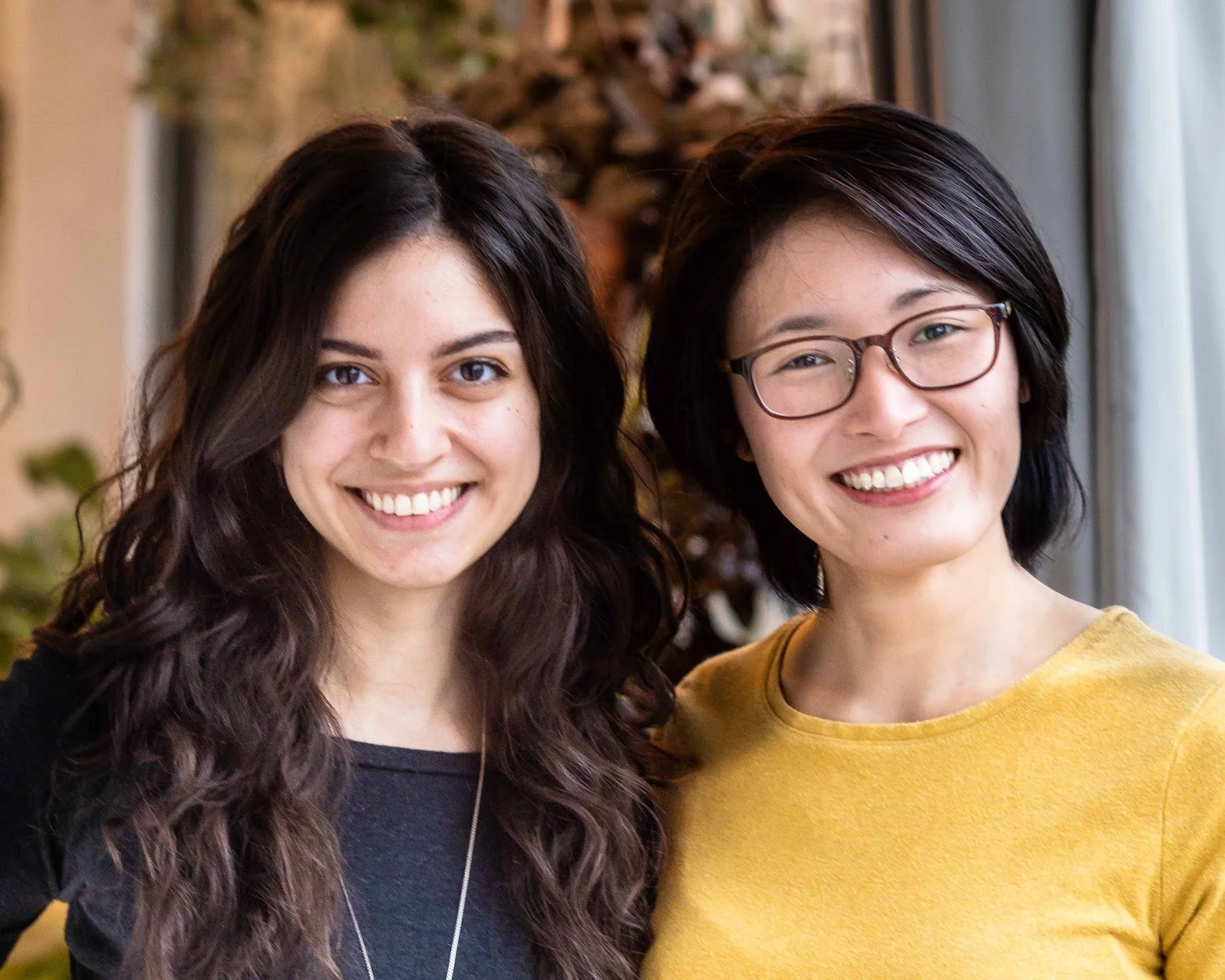Close-up of two women smiling, one with long dark wavy hair and the other with short dark hair wearing glasses, standing indoors with a Christmas tree in the background.