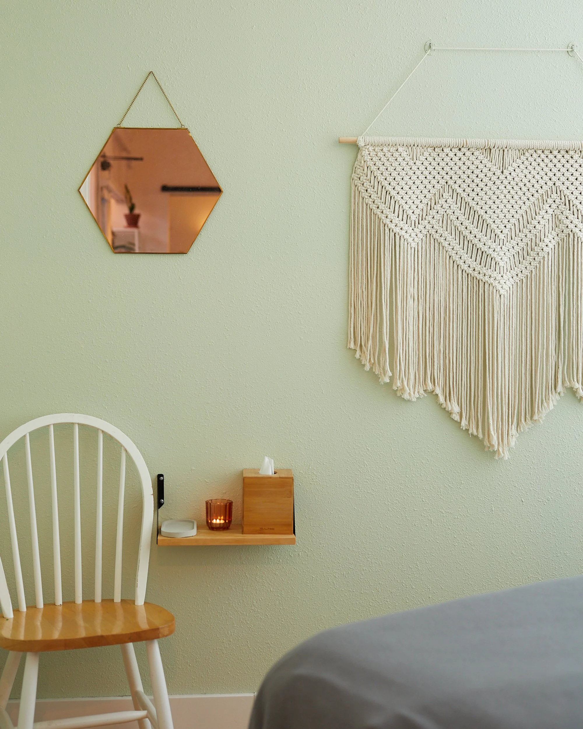 A cozy corner of a room with a white wooden chair, a small wooden wall shelf with a candle and tissue box, a decorative wall hanging made of woven material, a hexagonal mirror reflecting a potted plant, and a part of a gray bedspread in the foreground, all on a light green textured wall.