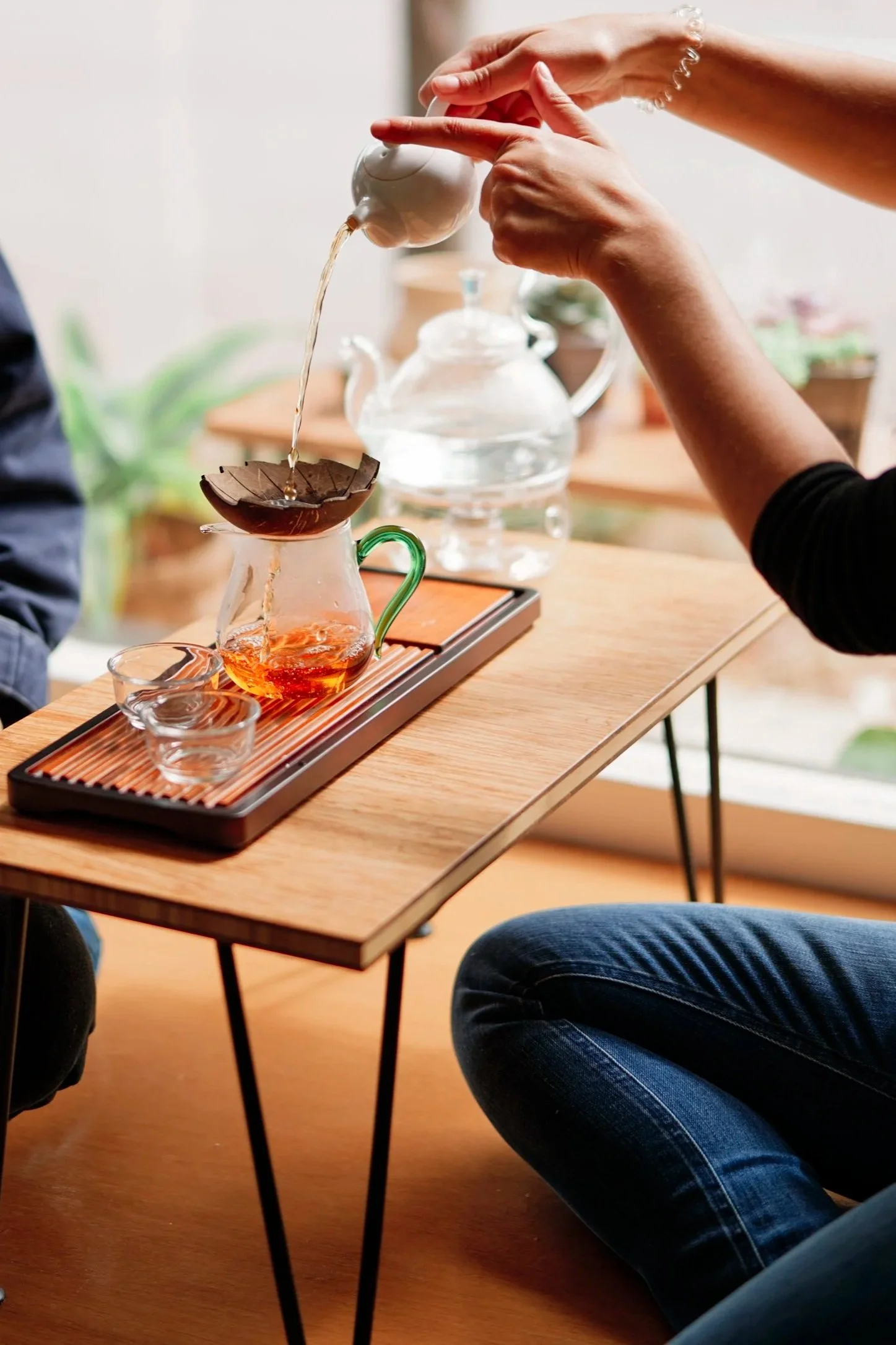 Person pouring hot water from a teapot into a glass tea cup on a wooden table. In the background, there is a glass teapot and other tea accessories.