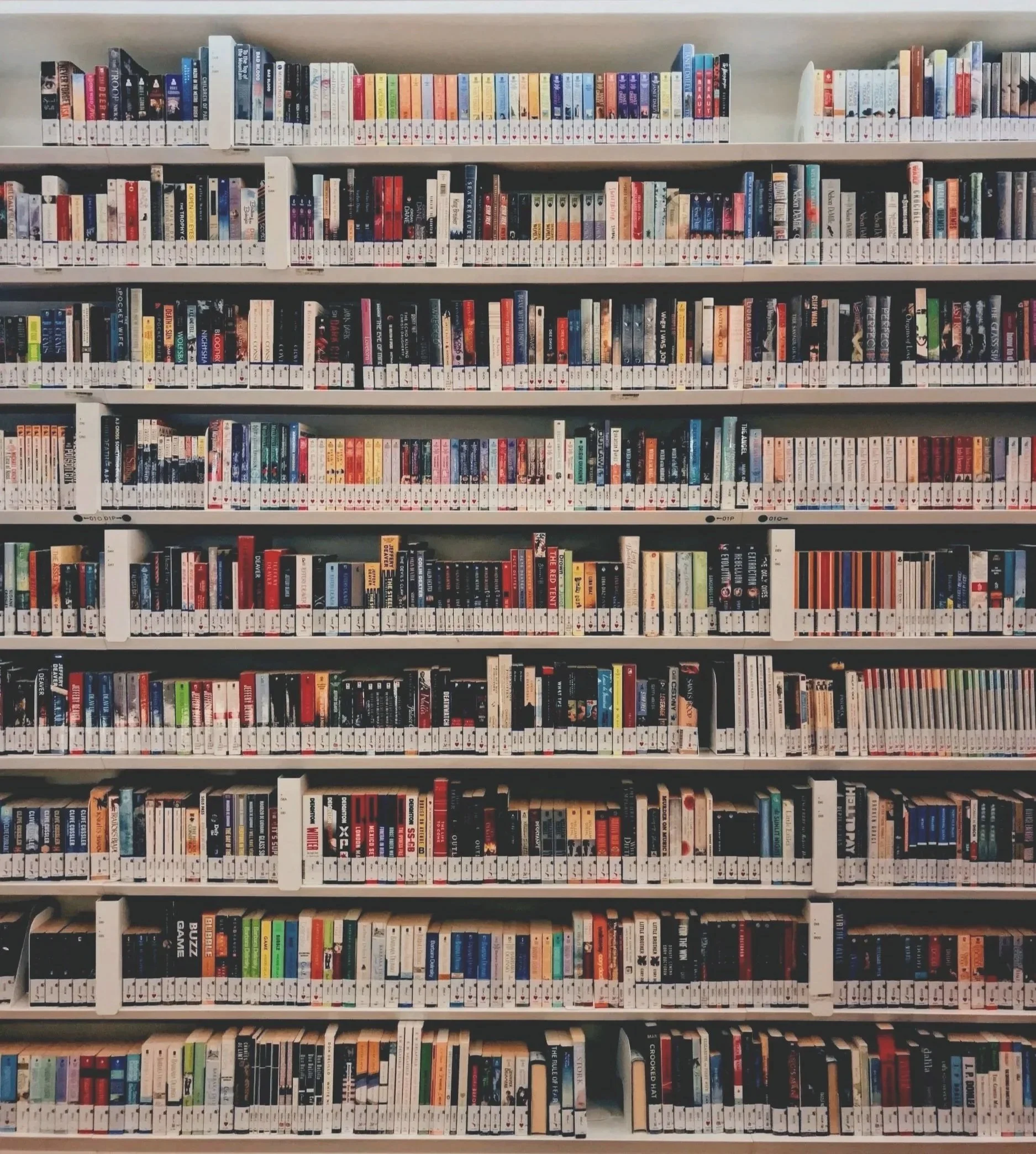 A photo of multiple white bookshelf shelves filled with a large collection of books, organized with white labels at the bottom of each book spine.