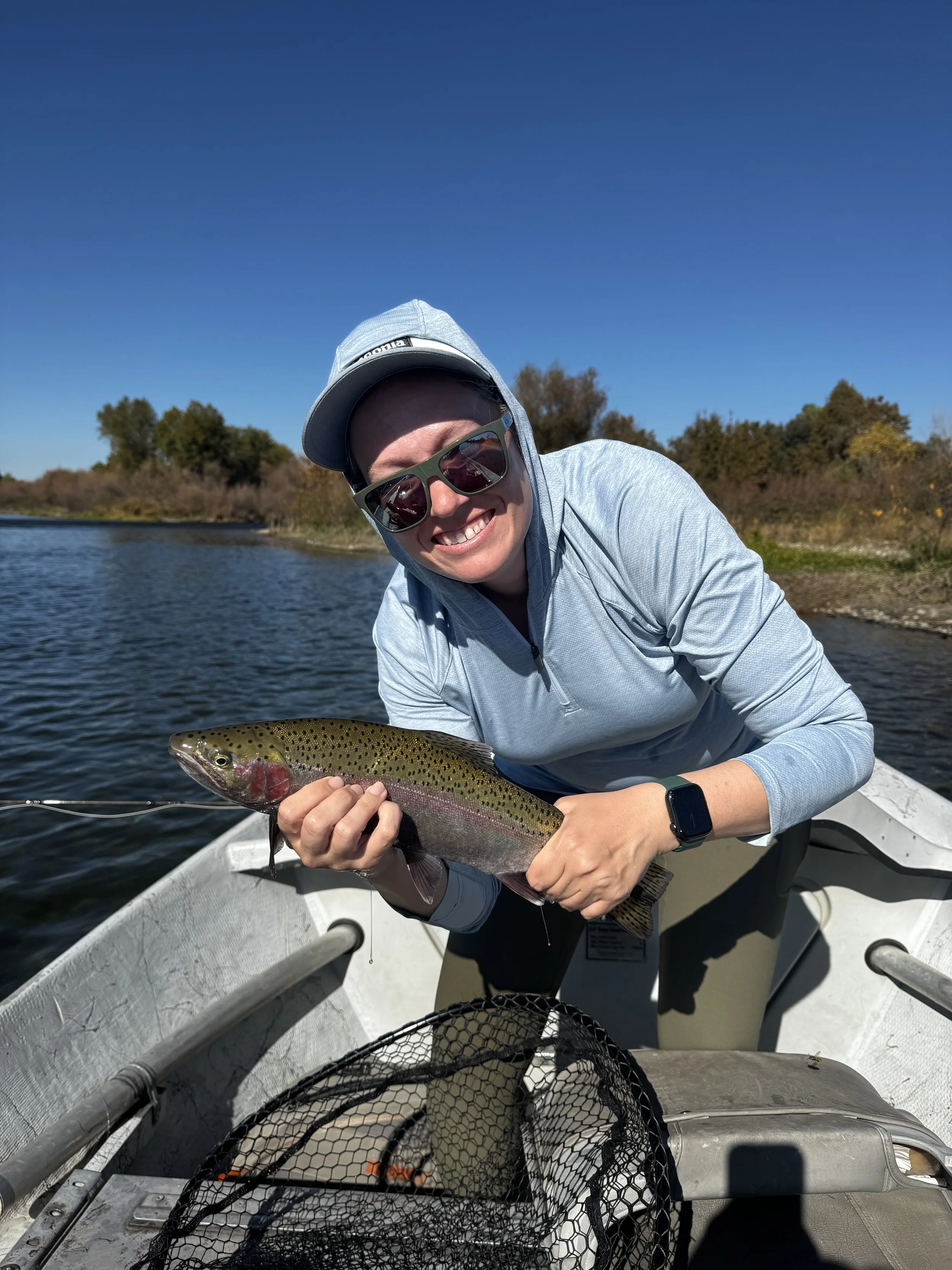 Person in a boat holding a rainbow trout caught while fishing, smiling and wearing sunglasses, a cap, and a hoodie, with trees and water in the background.