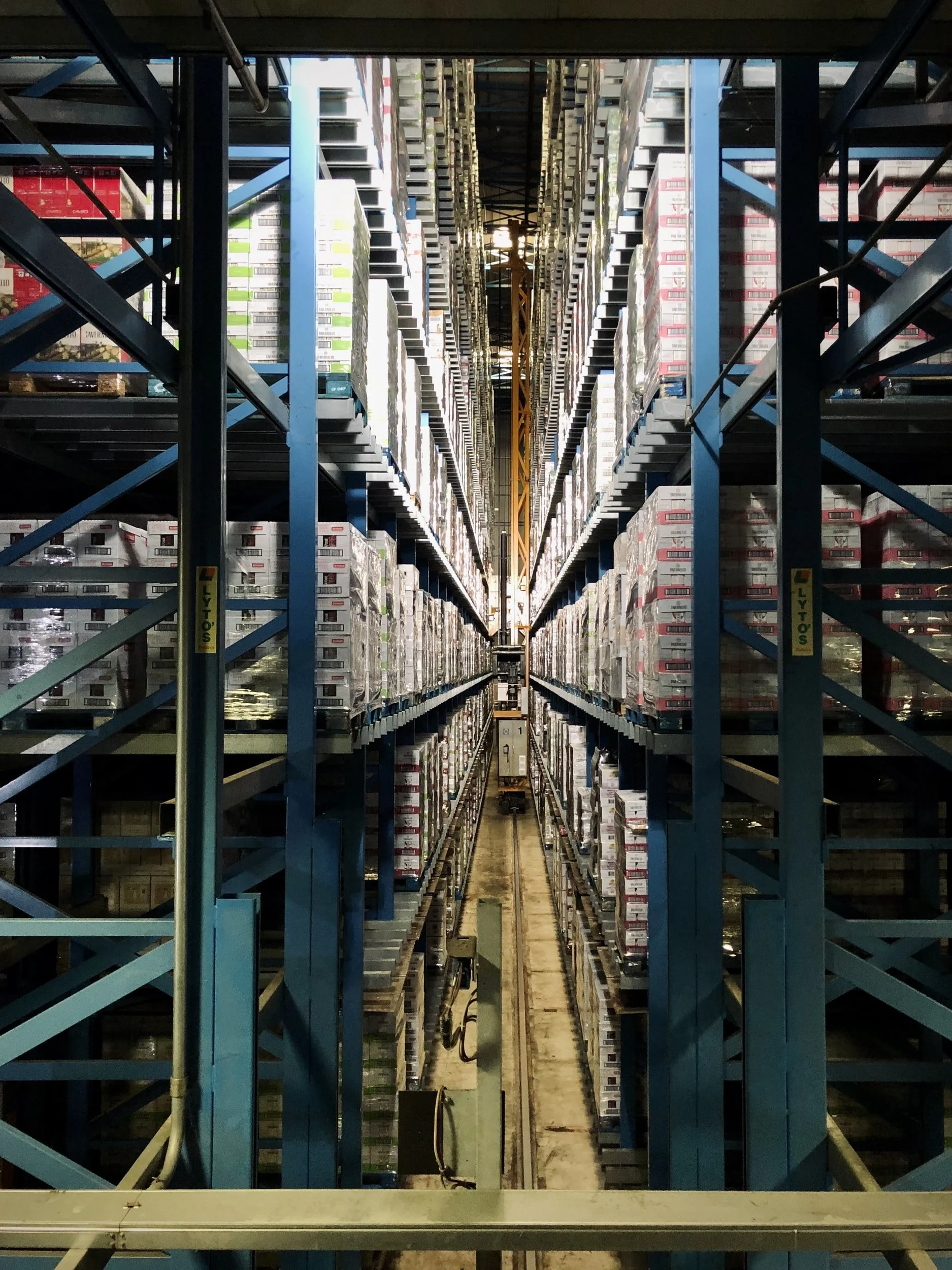 View of tall, industrial warehouse shelves filled with stacked boxes and pallets, with a narrow aisle in the center and a yellow crane lifting system overhead.