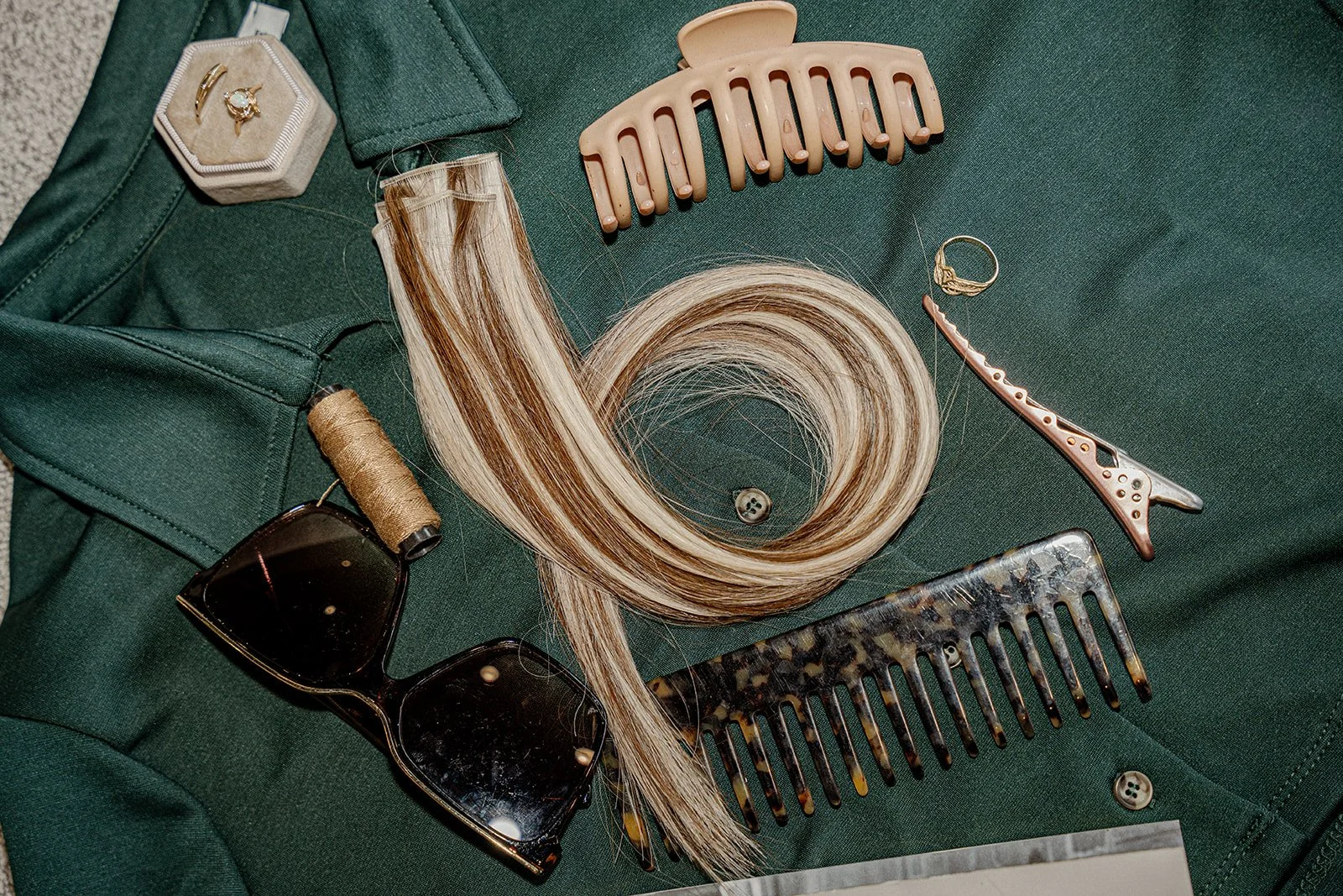A mix of items from someone's bag on a green shirt including hair extensions, sunglasses, combs, and claw clips.