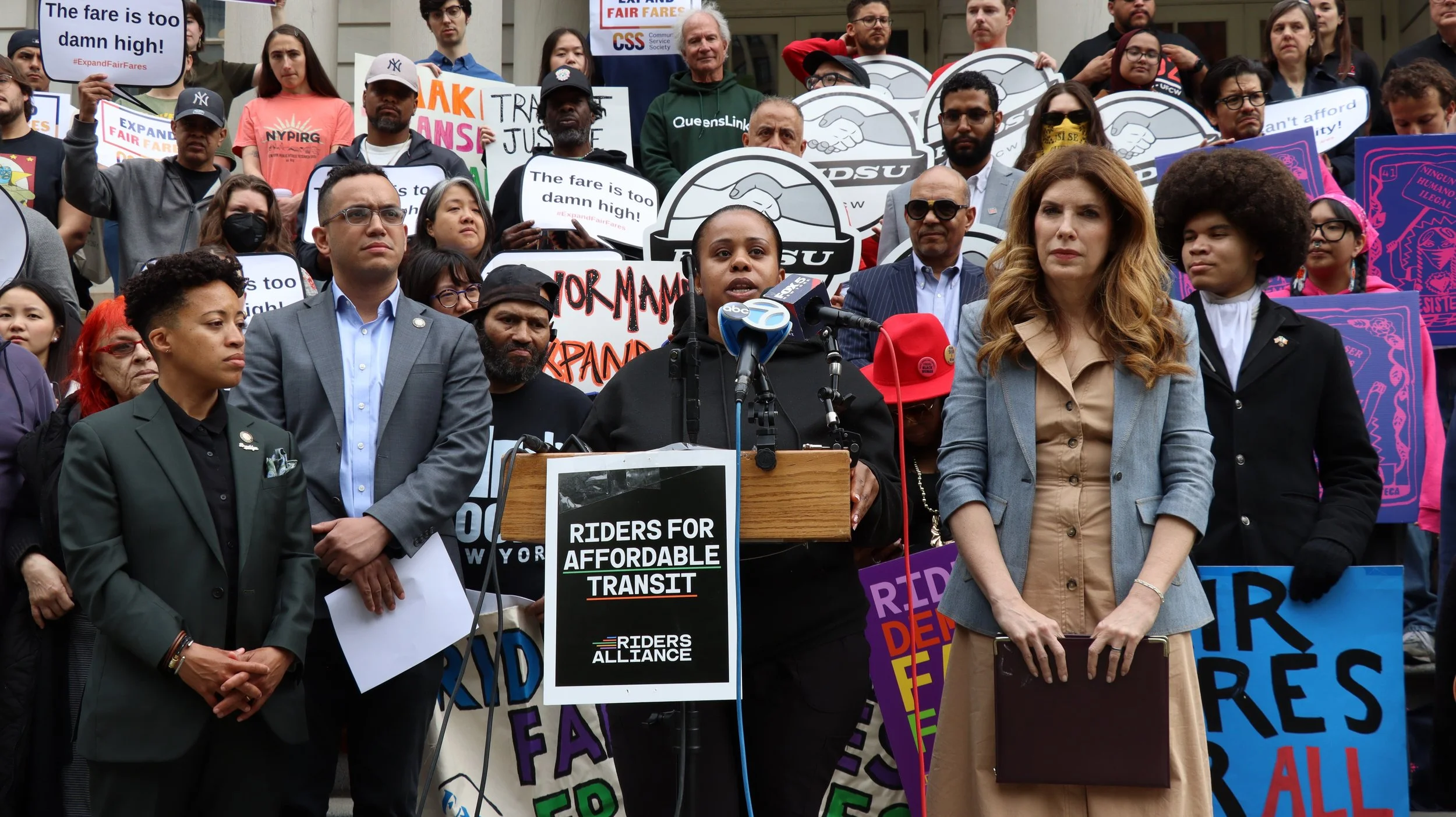 People rallying on the steps of New York City Hall with signs. A speaker is at a podium flanked by elected leaders and advocates. A black sign on the podium reads "Riders for Affordable Transit" and has a "Riders Alliance" logo at the bottom.