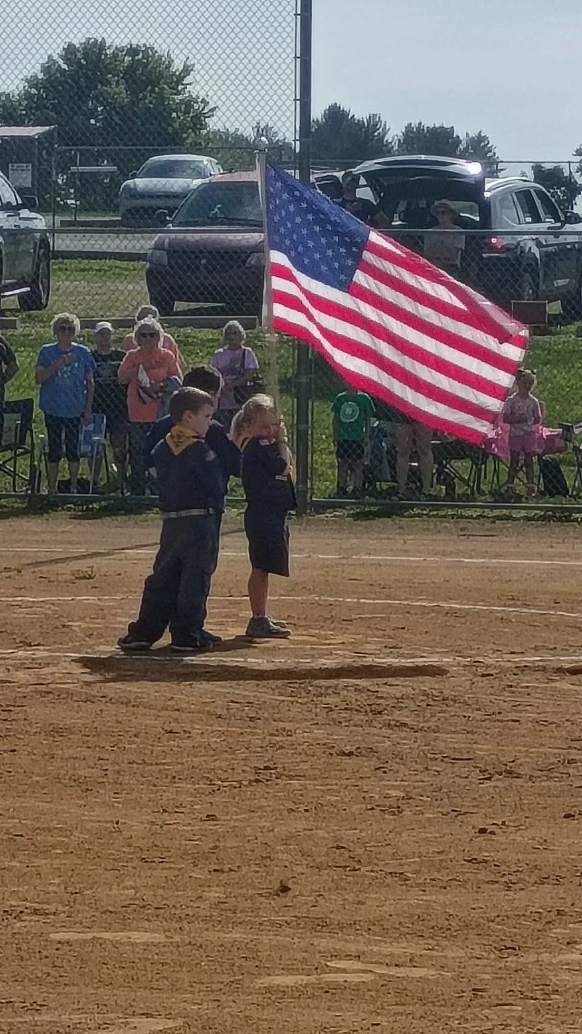**Alt text:** Two Cub Scouts stand on a baseball field holding a large American flag during a ceremony. Spectators watch from behind a chain-link fence, with parked vehicles and trees in the background.