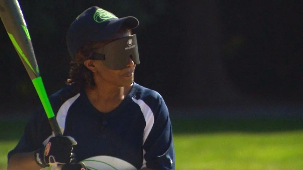 A young person wearing a dark sports shirt, black gloves, and a cap, with eye covering, holding a green and black bat, outdoors on a grassy field during daytime.