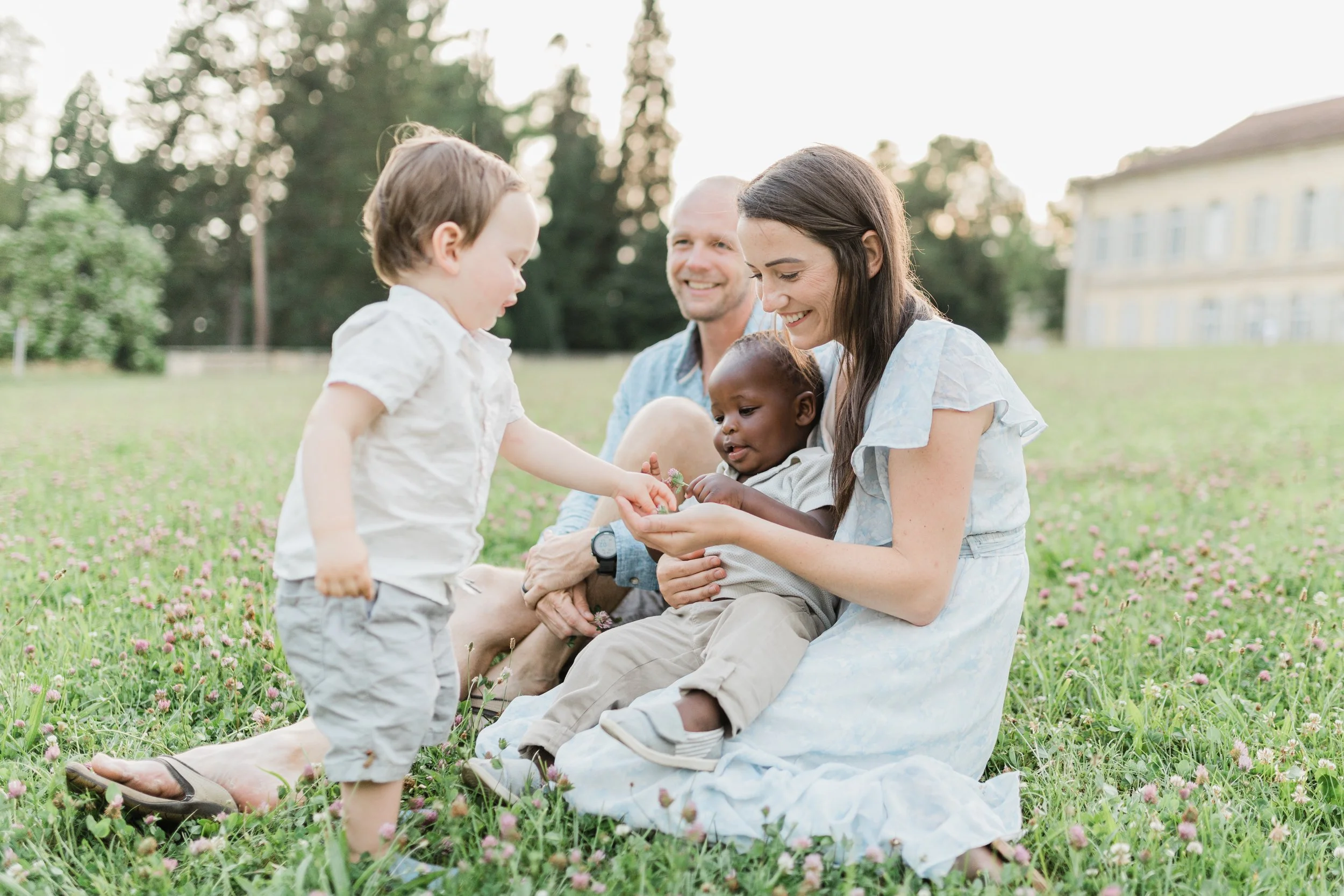 A family of four enjoying time together outdoors on a grassy field with pink flowers, with trees and a building in the background.