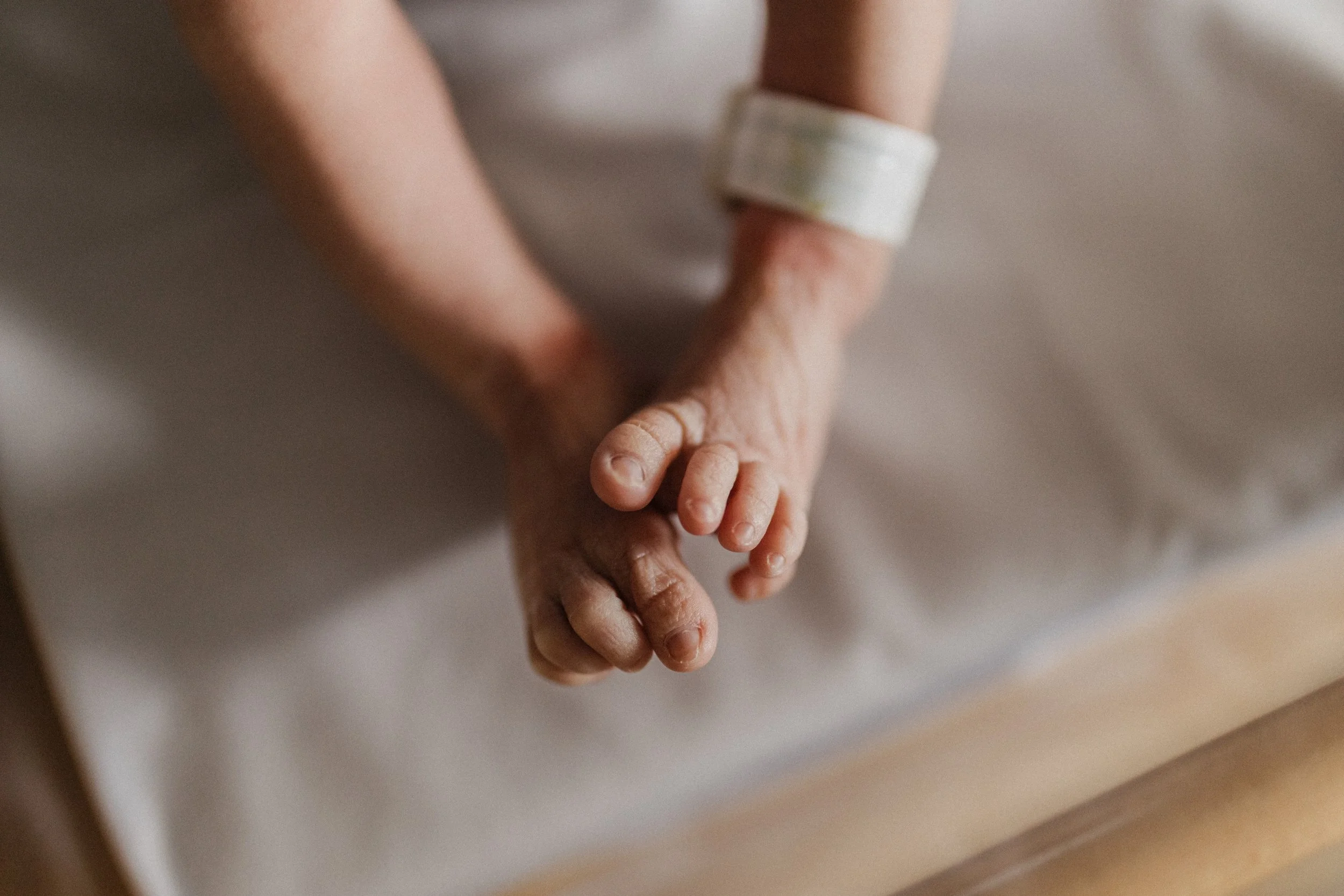 Close-up of elderly person's hands resting on a bed, one hand clasping the other, wearing a hospital wristband.
