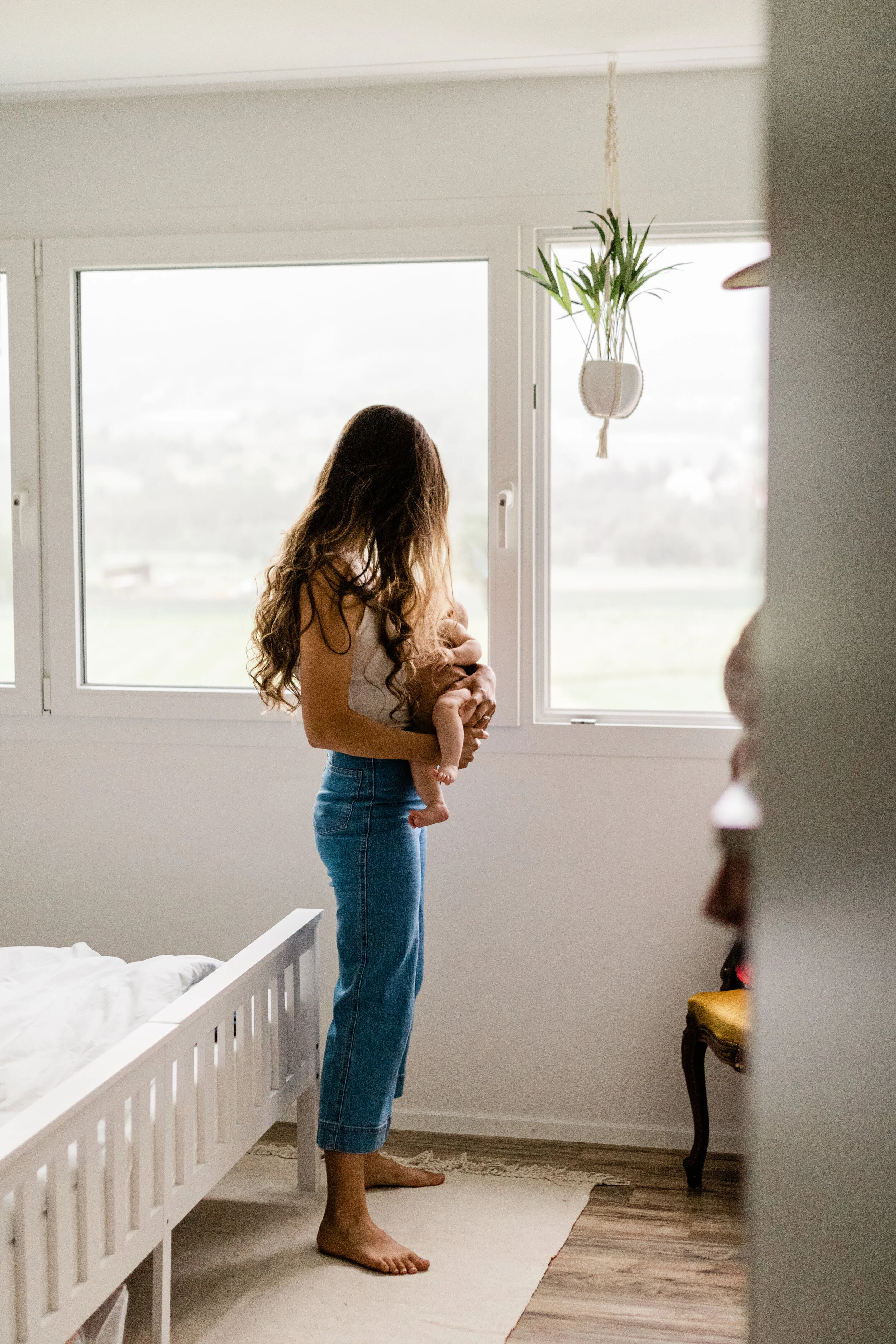 A woman standing by a window holding a baby in her arms, looking outside in a bright, minimalistic bedroom with white walls and wooden flooring.
