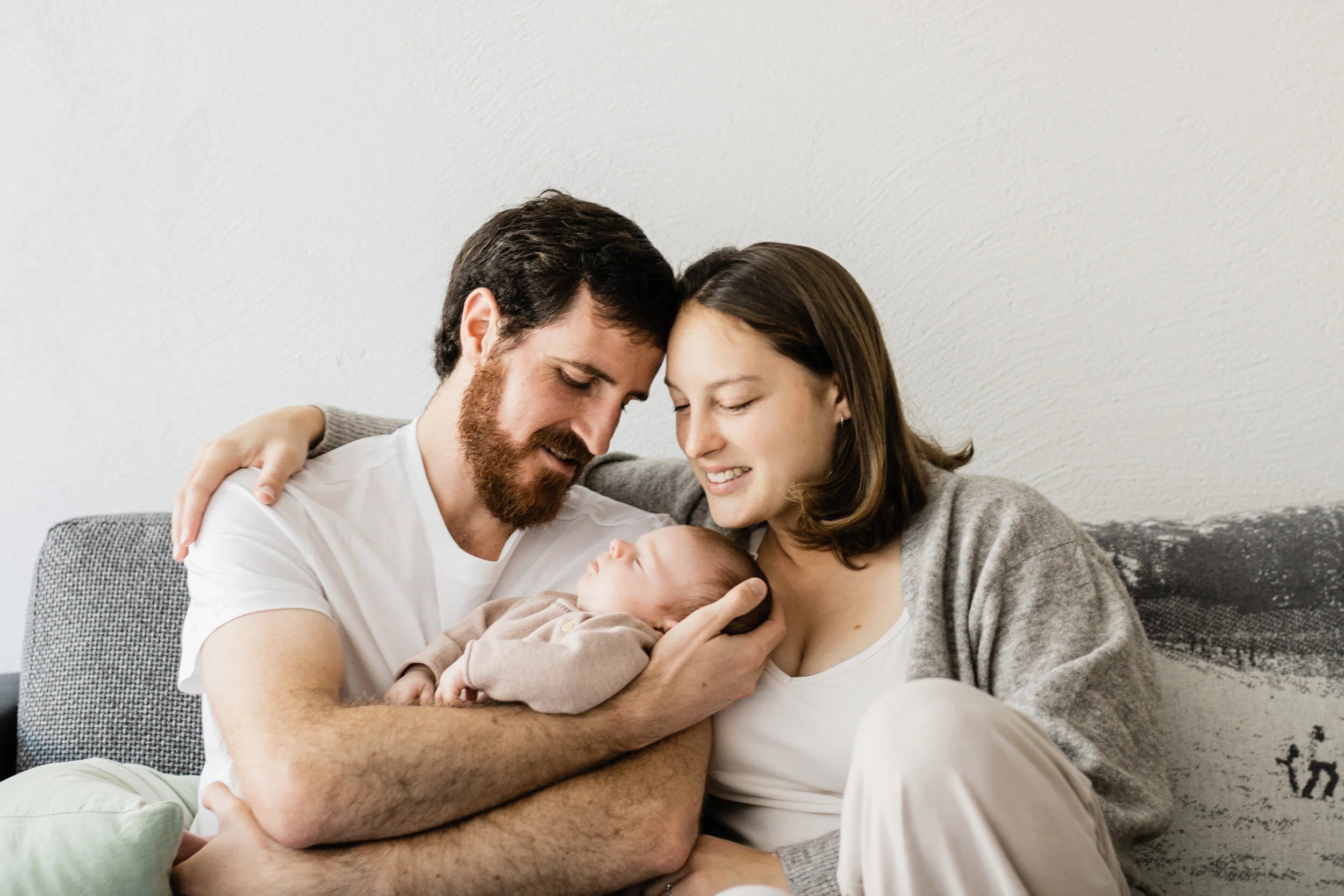 A happy family of three, parents with their newborn baby, sitting on a couch indoors, embracing and smiling at the baby.