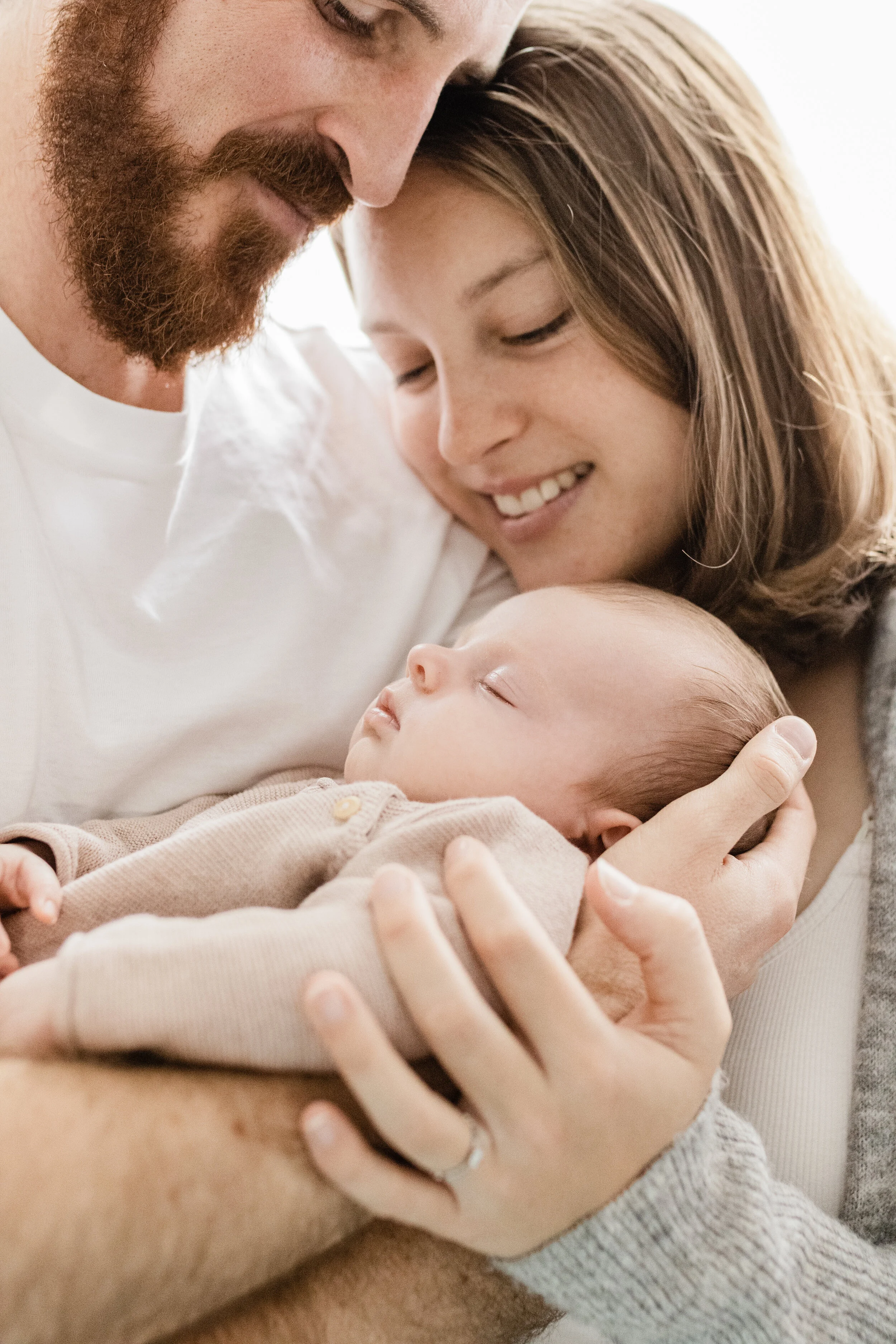 Family of three, father, mother, and sleeping baby, sharing a tender moment indoors.