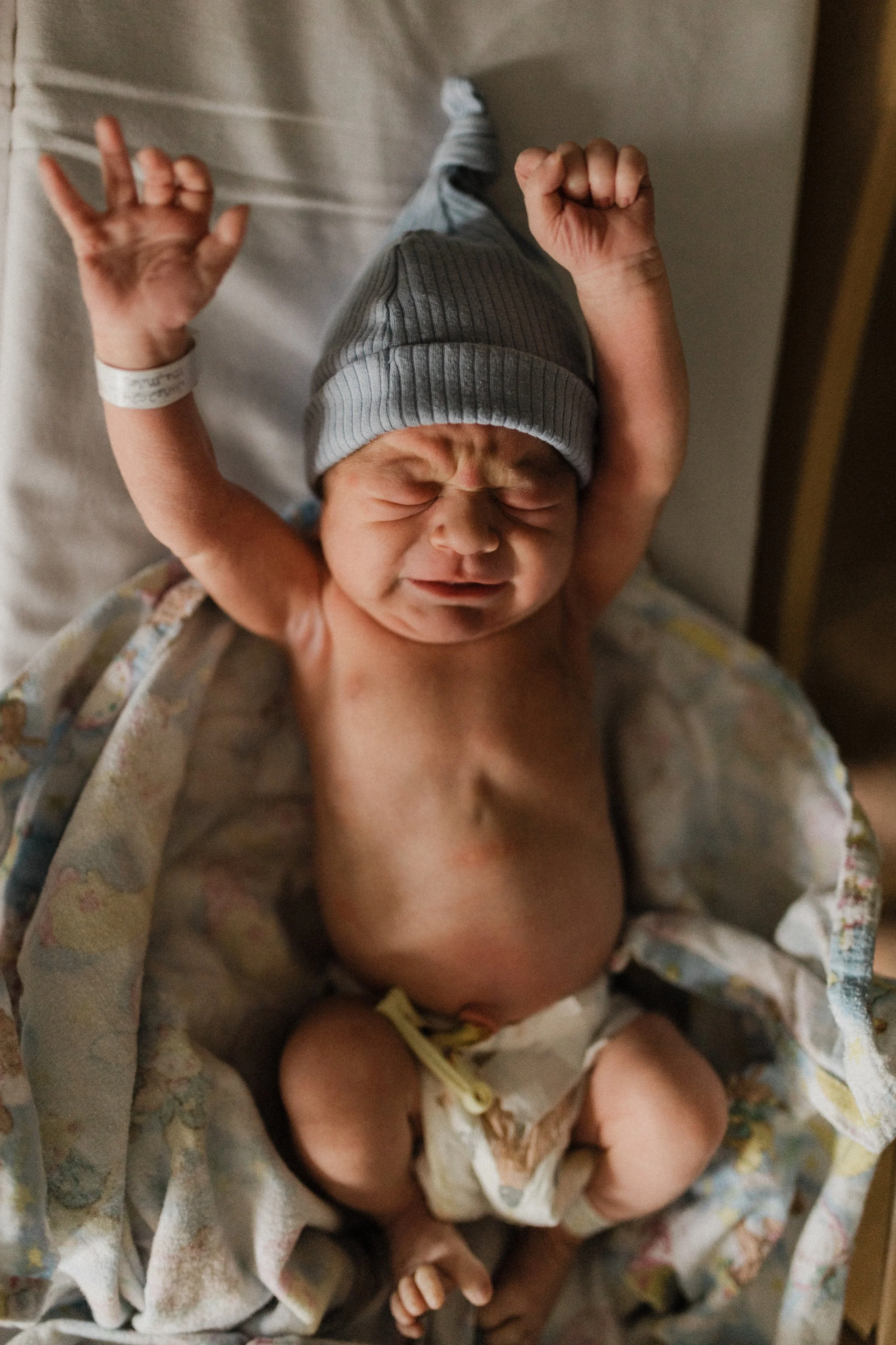 Newborn baby with arms raised, wearing a gray beanie, lying on a bed with a hospital wristband, wrapped in a floral blanket, crying with eyes shut.