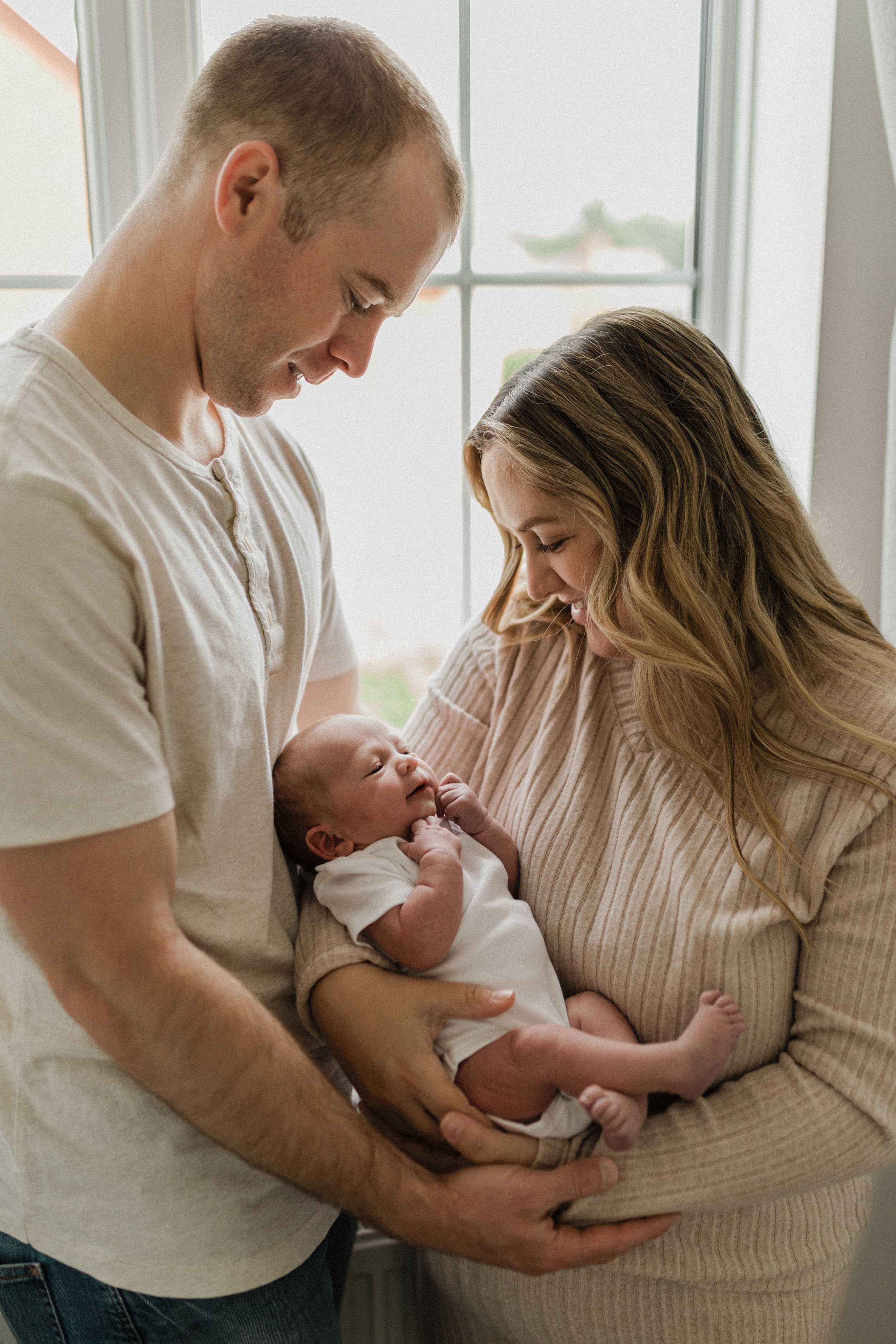A couple holding their newborn baby by a window, smiling and looking at the infant.