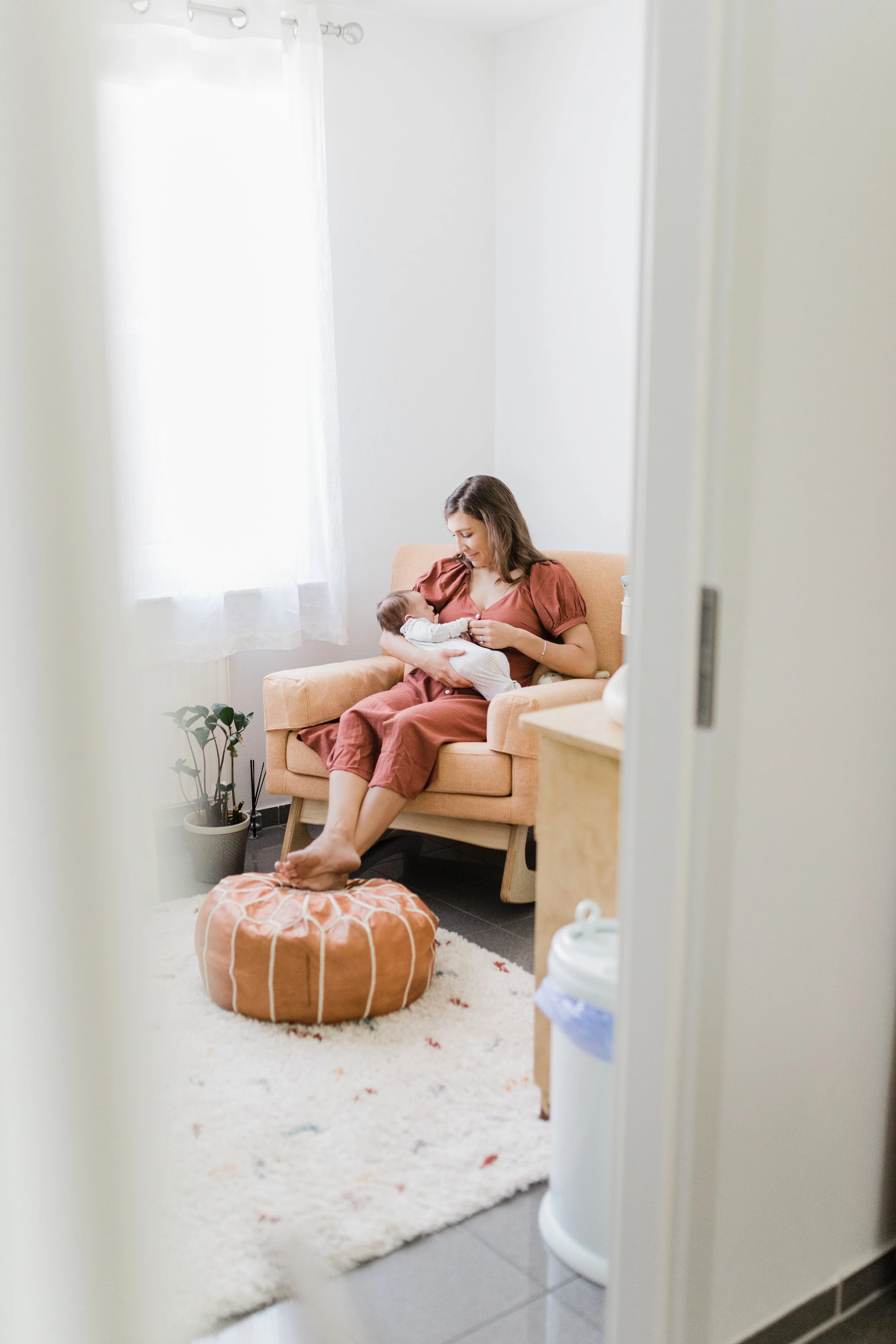 A woman sitting on a beige armchair breastfeeding a baby in a room with white walls, a window with white curtains, a potted plant, a colorful rug, and a pouf, seen through a partially opened door.
