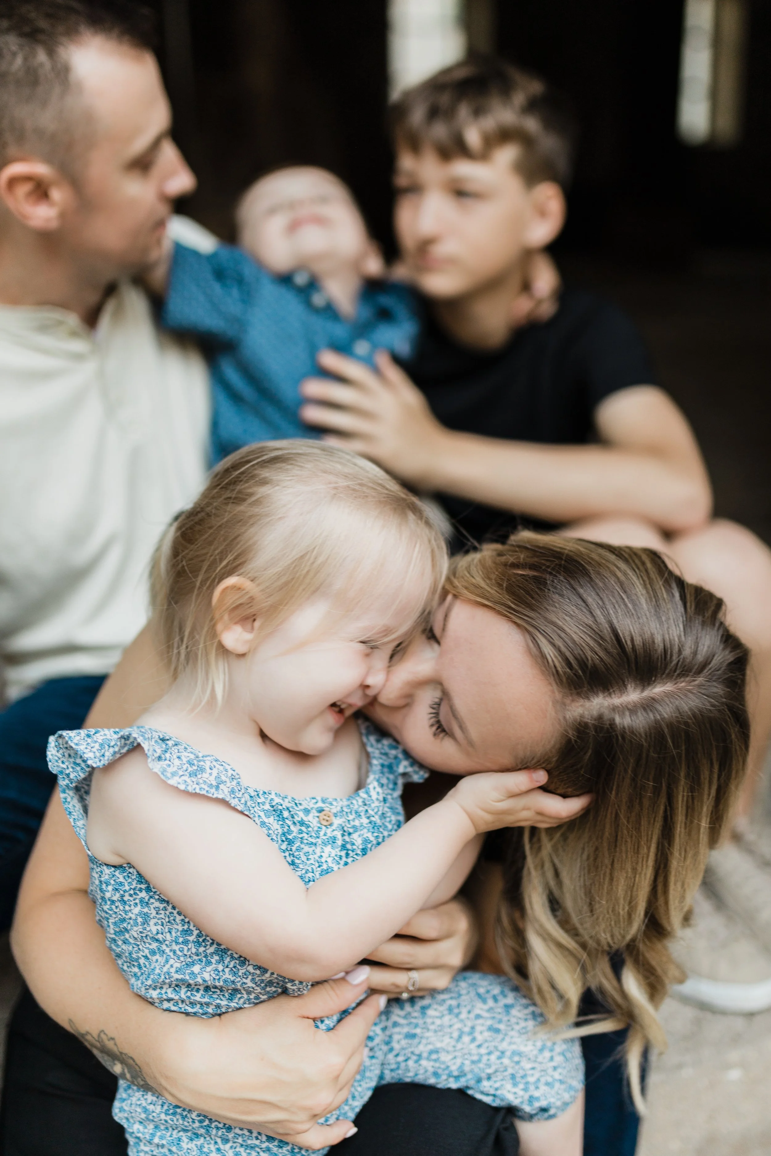 A family of six affectionately interacting, with a woman holding a young girl and a man, an older boy, and two younger boys in the background.