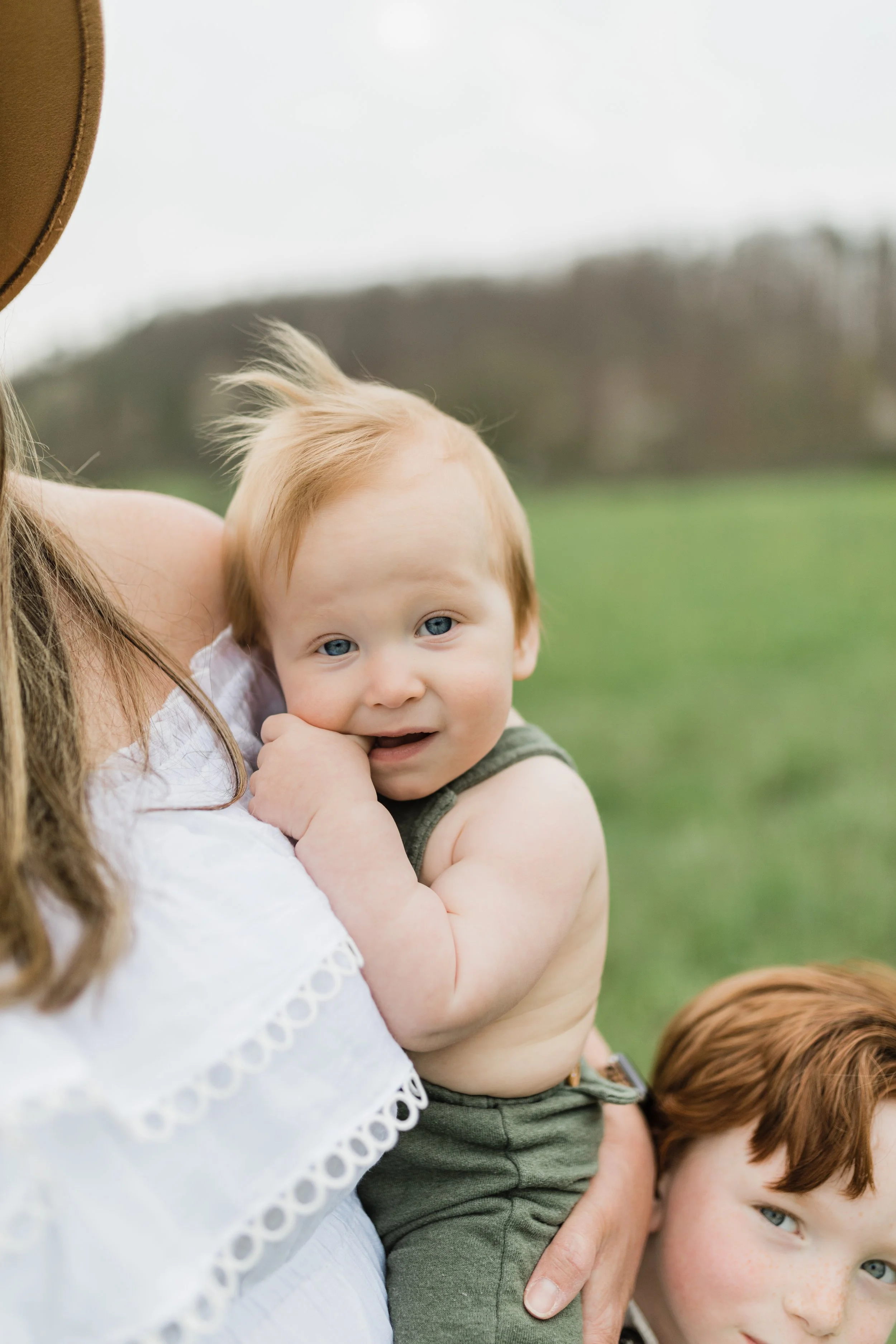A toddler with blue eyes and red hair resting his head on a woman's shoulder outdoors.