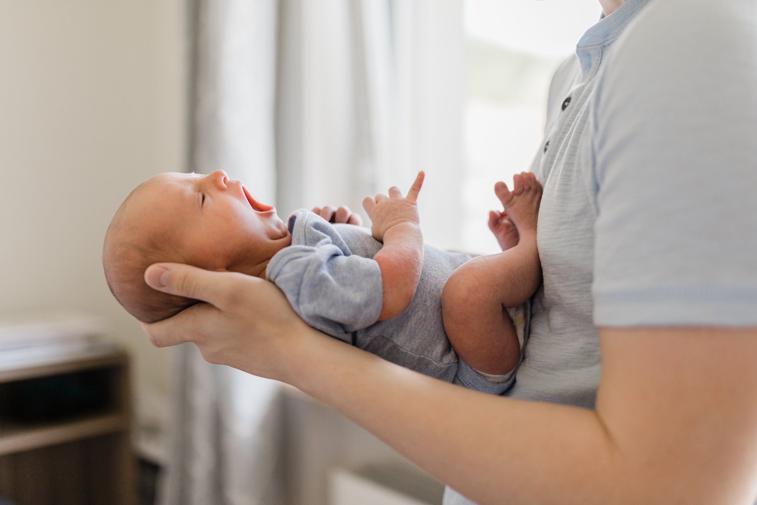 A person holding a yawning baby close to their chest indoors, with a window and curtains in the background.