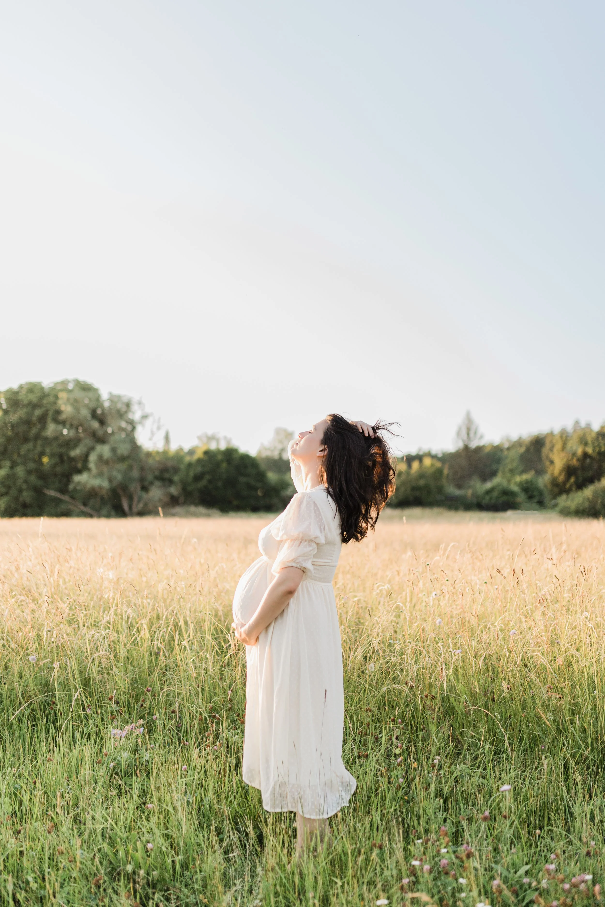 Pregnant woman in a white dress standing in a field of tall grass, facing the sky with eyes closed and hair lifted by the breeze, during sunset.