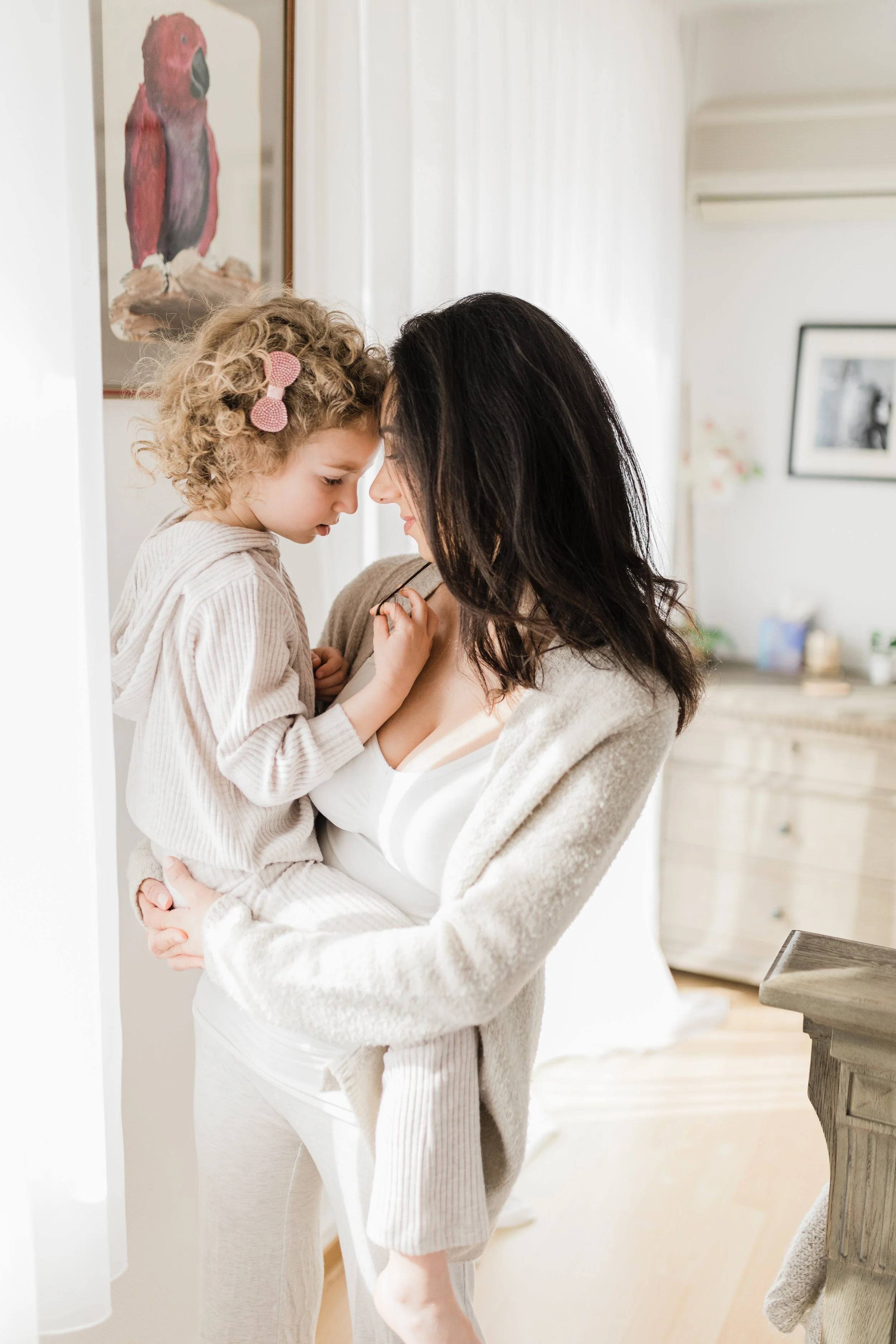 A woman and a young girl with curly blonde hair and a pink hair clip share a tender moment, touching foreheads and smiling in a bright room.