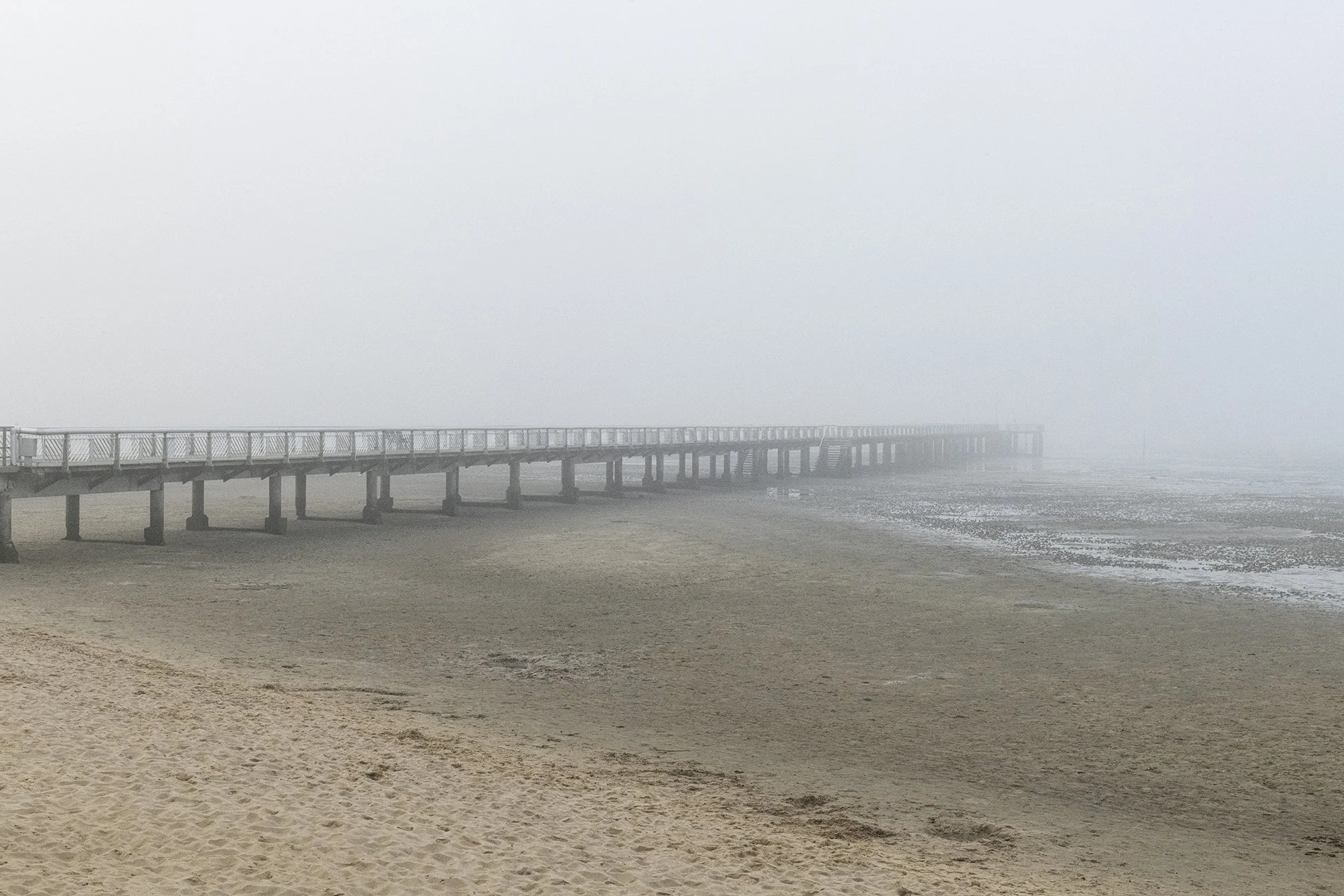Ponton s'étendant sur une plage brumeuse, sous un ciel nuageux.