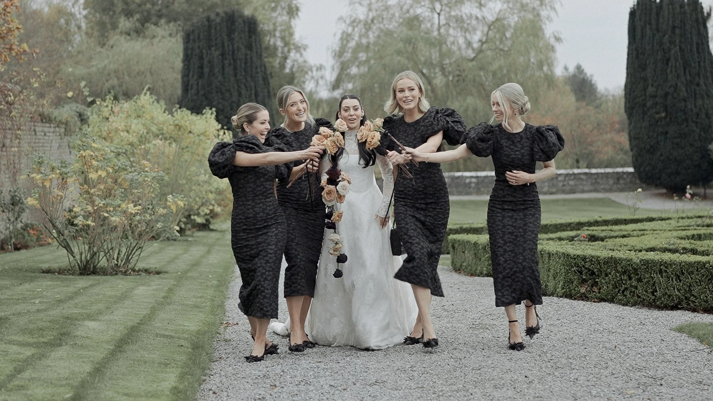 Bride and bridesmaids in black dresses walking through the gardens at Kilkea Castle