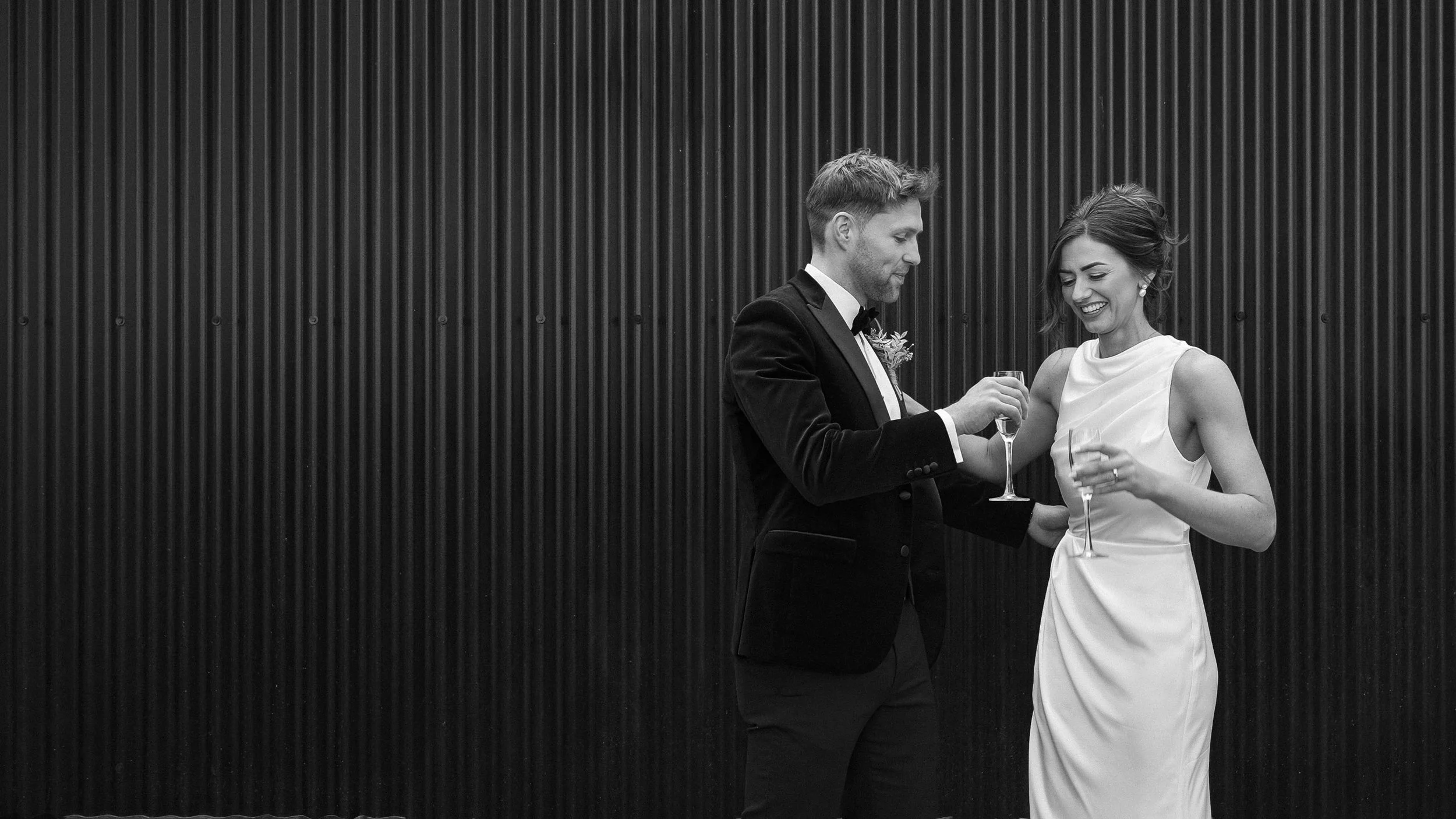 Newlyweds laughing with glasses of bubbles against a black wall at Barberstown Castle