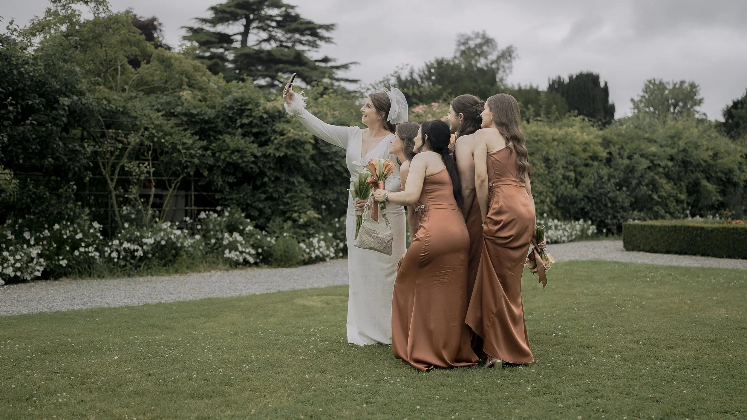 Bride taking a selfie with her bridesmaids in copper satin dresses in the gardens of Kilkea Castle