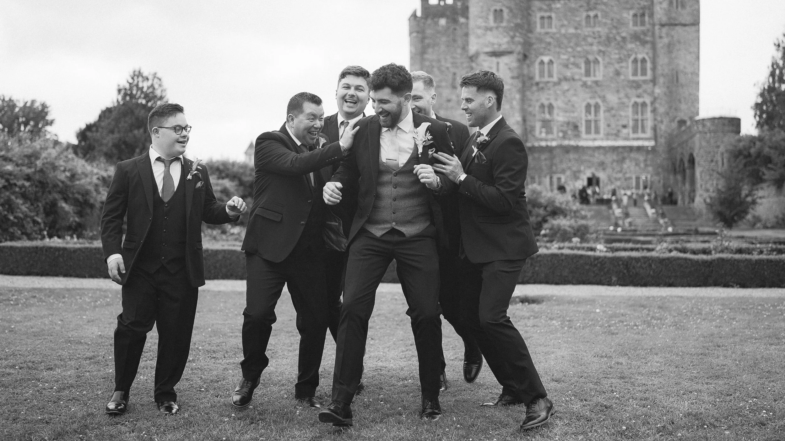 Laughing groomsmen lifting the groom on the lawn in front of Kilkea Castle, black and white