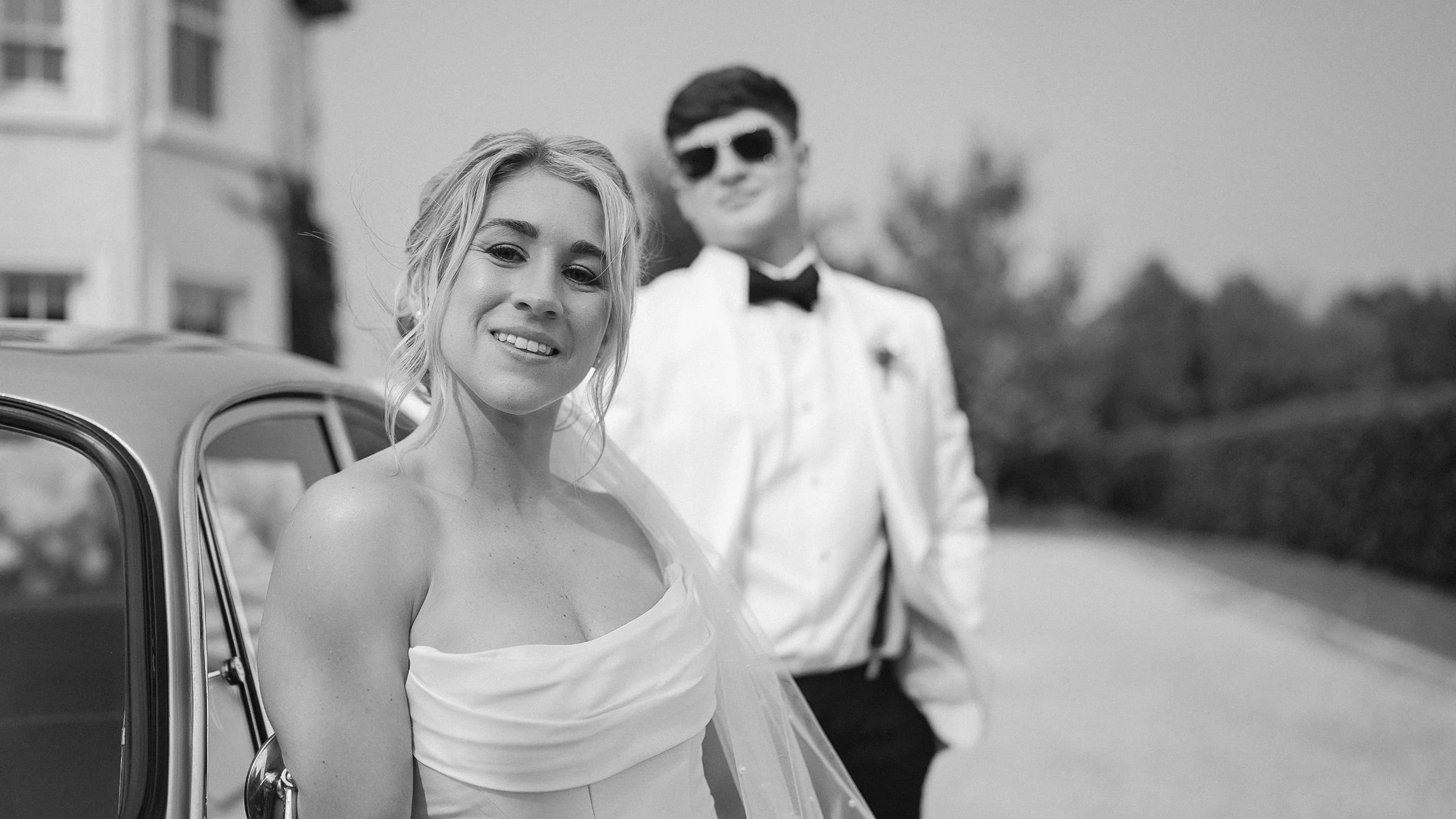 Bride smiling by a vintage wedding car with the groom behind her – Curragh wedding, Kildare