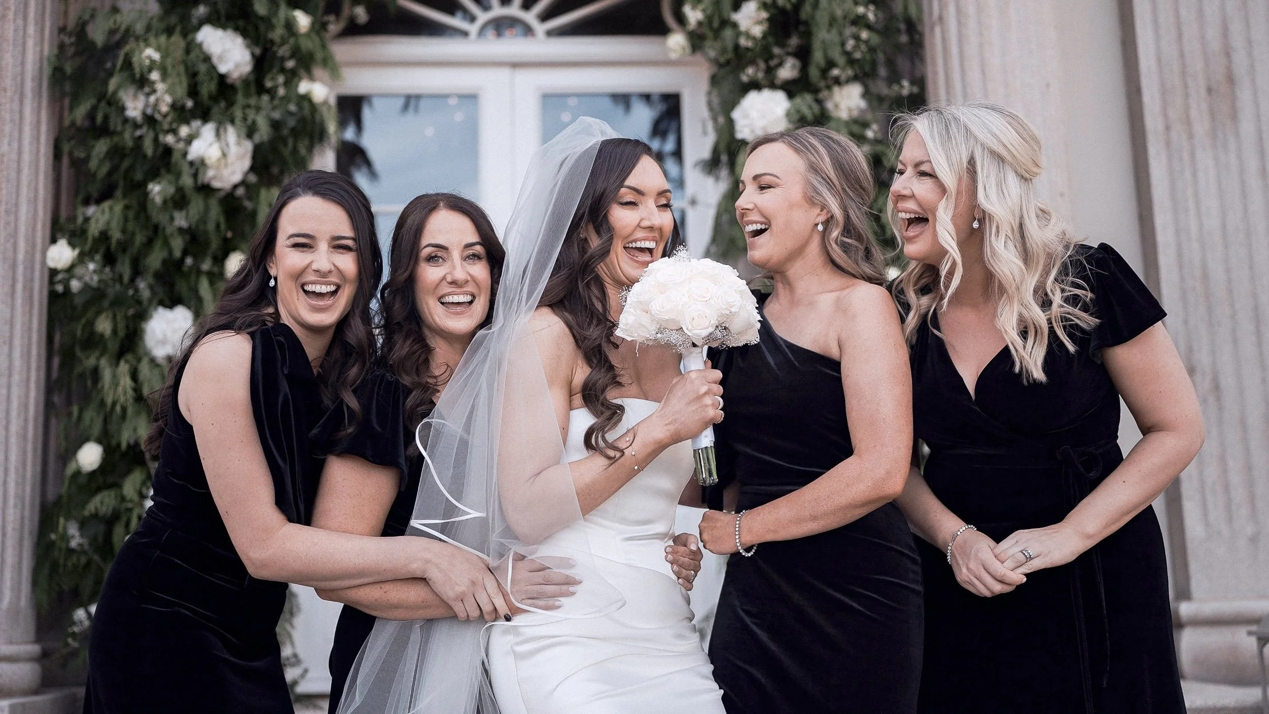 Bride laughing with her bridesmaids in black velvet dresses at Clonwilliam House, Co. Wicklow