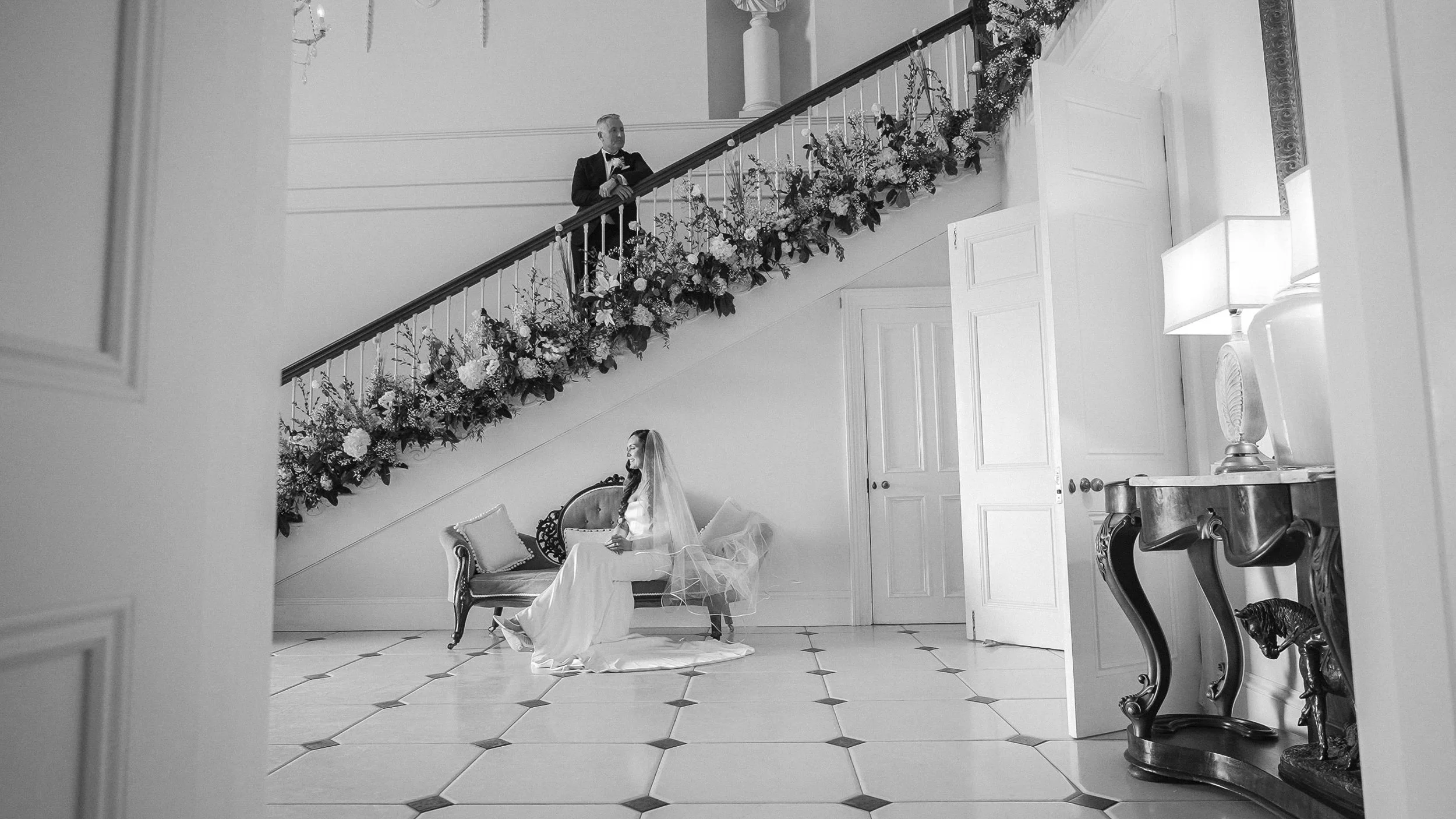 Black and white photo of the groom on the flower-dressed staircase and the bride below in the hall of Clonwilliam House