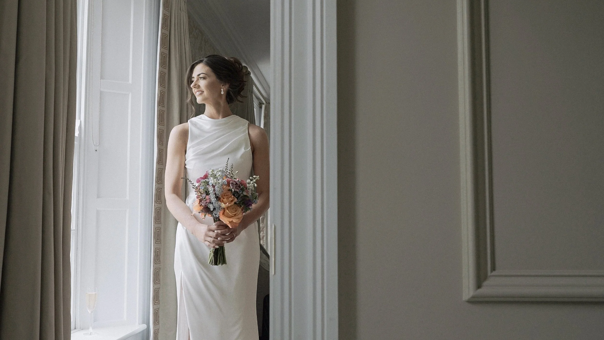 Bride in a minimal satin gown holding a colourful bouquet by the window at Barberstown Castle, Co. Kildare