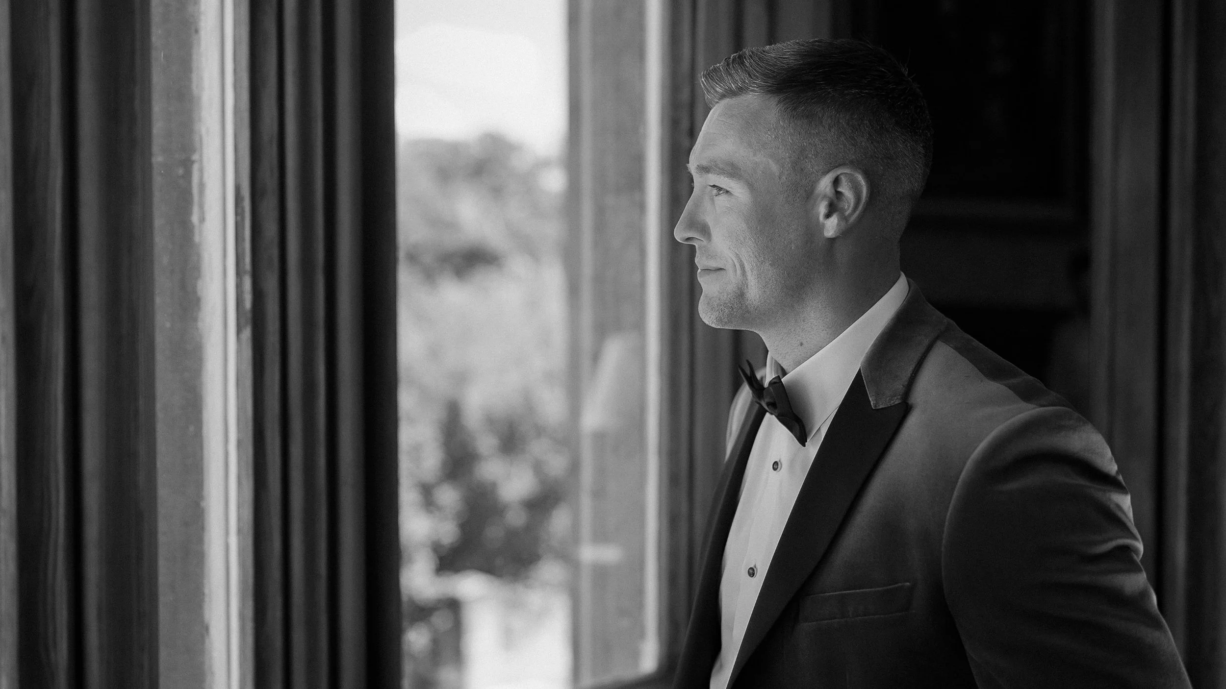 Groom in a velvet tuxedo jacket and bow tie looking out of a window at Markree Castle, black and white